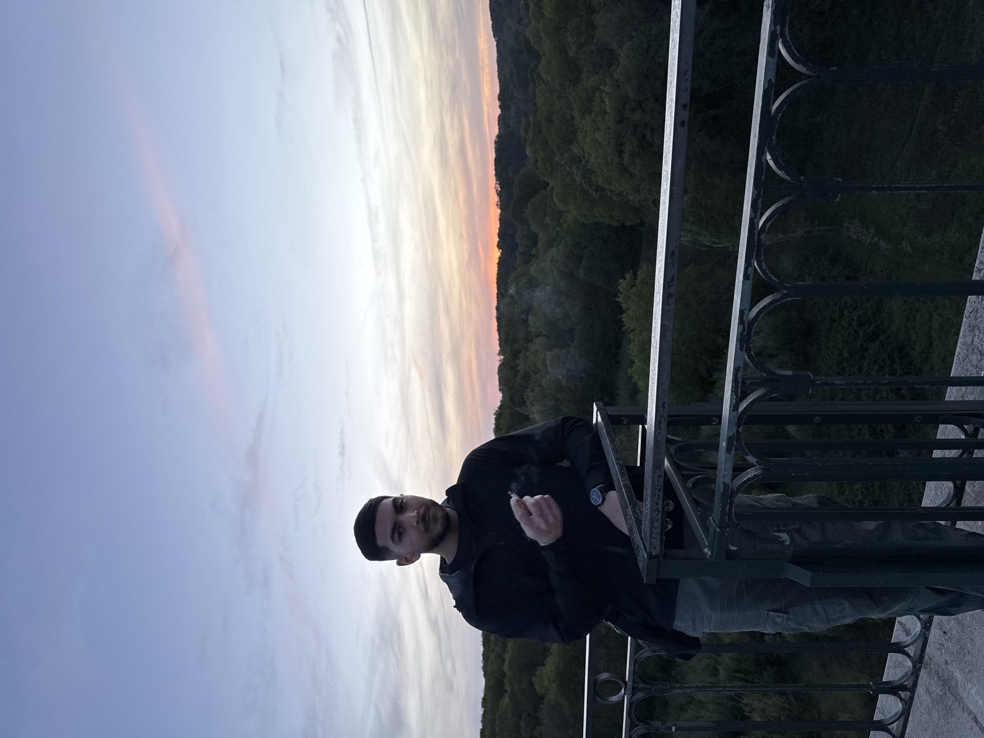 A portrait-oriented, eye-level shot captures a young man outdoors at sunset, in Gometz-le-Châtel, France. The man, with short dark hair and a beard, is positioned slightly to the left of the frame's center. He wears a dark long-sleeved top and dark pants, and a watch on his left wrist. His right hand holds a lit object, possibly a cigarette or joint, from which smoke trails upwards. He leans casually against a dark, ornate metal railing that forms the lower-right boundary of the image.
The background features a dramatic sky with soft, elongated clouds displaying warm hues of orange, pink, and pale yellow near the horizon, transitioning to light blue and grey further up. Below the horizon, a dense, dark green forest stretches across the landscape, suggesting a valley or a view from a high vantage point. The overall lighting is dim, characteristic of twilight. The man looks directly at the camera with a neutral expression. No visible text is present in the image.
