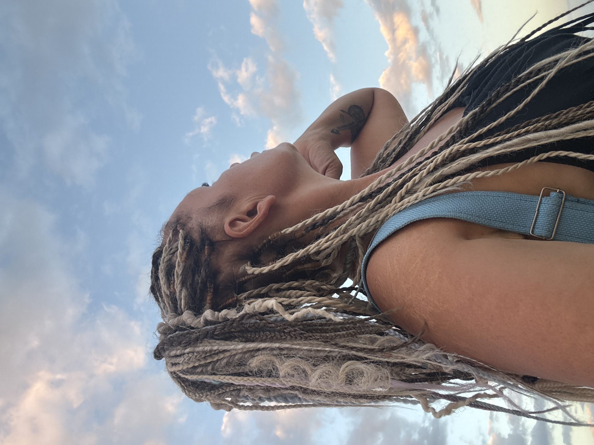 A woman with braided hair is looking up at the sky. She is wearing a black top and blue overalls. The sky is blue with some clouds. The image appears to have been taken outdoors. The location is Kuta, Indonesia.