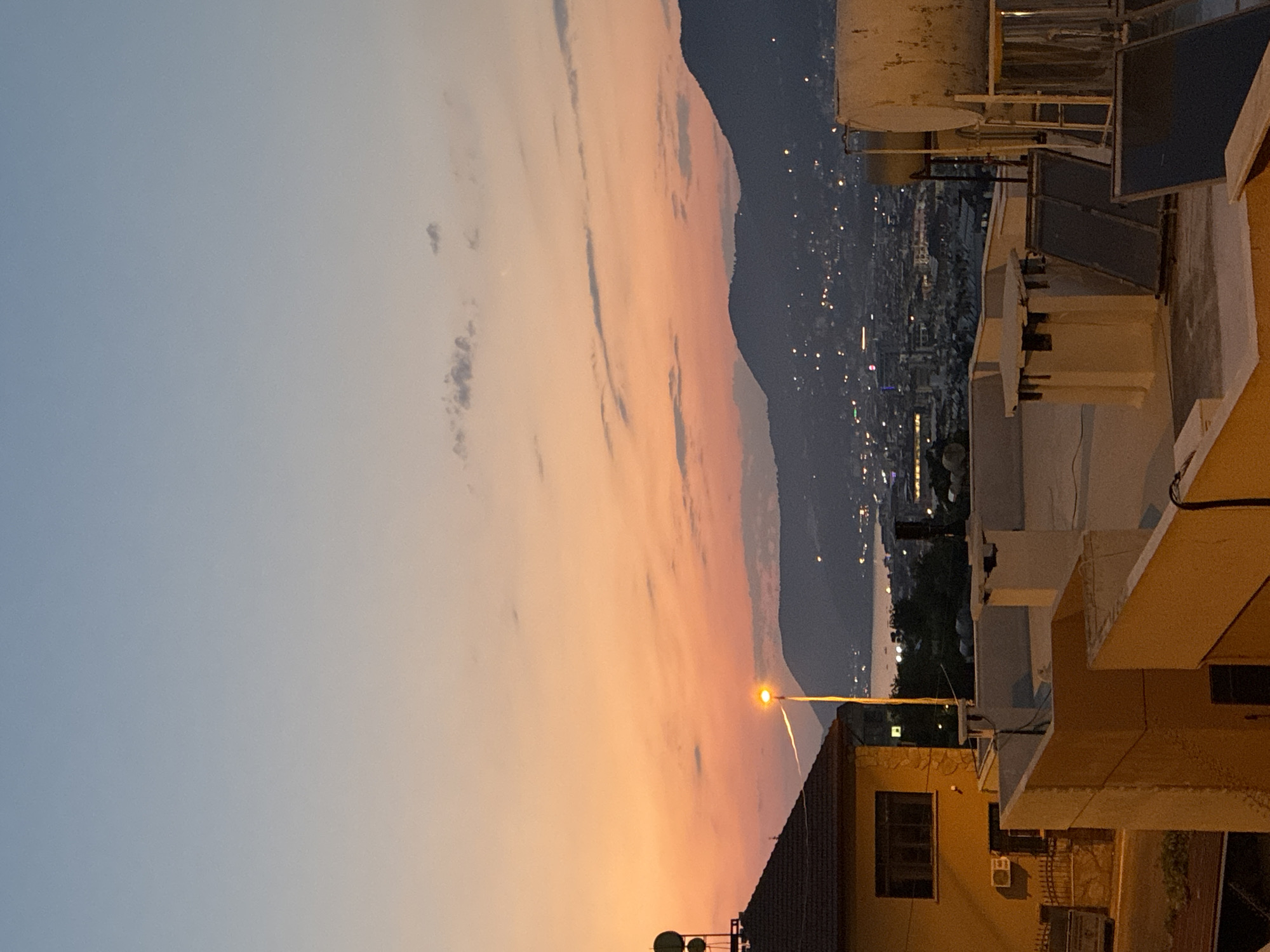 The image captures a twilight scene in Alanya, Turkey, overlooking a city nestled at the foot of dark, imposing mountains. The sky is a gradient of soft blues and oranges, with wispy clouds scattered across it. In the foreground, the rooftops of several buildings are visible. One prominent building on the left is painted yellow with a dark-tiled roof, featuring a few antennas on top. A streetlamp with a bright orange glow stands near it, casting a warm light.
The middle ground reveals the urban landscape of Alanya, with countless lights twinkling from buildings and along the coast. A body of water, likely the Mediterranean Sea, is visible between the city and the mountains. The mountains themselves are silhouetted against the darkening sky, their slopes dotted with more lights, suggesting habitation extending upwards.
The foreground also shows parts of other rooftops, including what appear to be solar panels and water tanks, common fixtures on buildings in sunny climates. The overall impression is one of a serene evening in a coastal city, with the natural beauty of the landscape meeting the man-made environment. There are no people visible in the image.