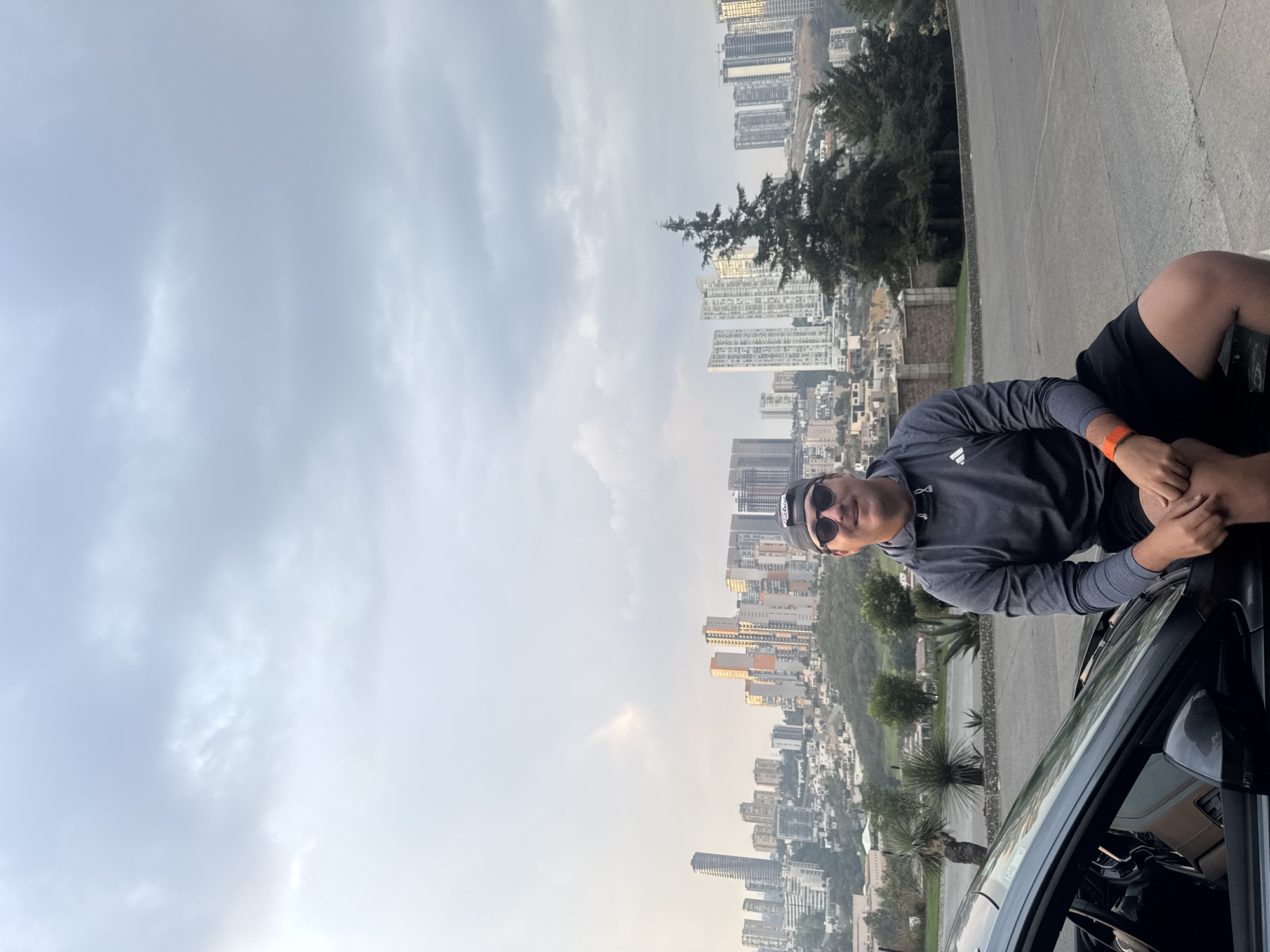 A man wearing sunglasses, a gray Adidas sweatshirt, a baseball cap, and an orange fitness band is sitting on the passenger side of a black car. The man is positioned against a backdrop of a city skyline in Huixquilucan de Degollado, Mexico. Numerous high-rise buildings are visible against an overcast sky. Trees and greenery are present among the urban structures.