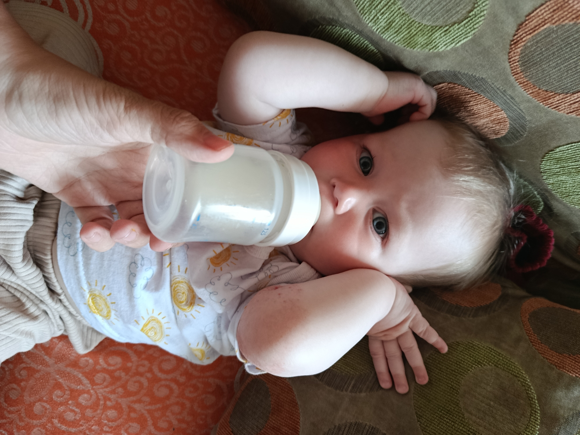 A fair-skinned infant with blue eyes is lying on a patterned orange and green pillow, being bottle-fed. An adult hand is holding the bottle. The infant is wearing a white shirt with sun designs and beige pants. The location in the city of Фоки, Russia is not discernible from the image.