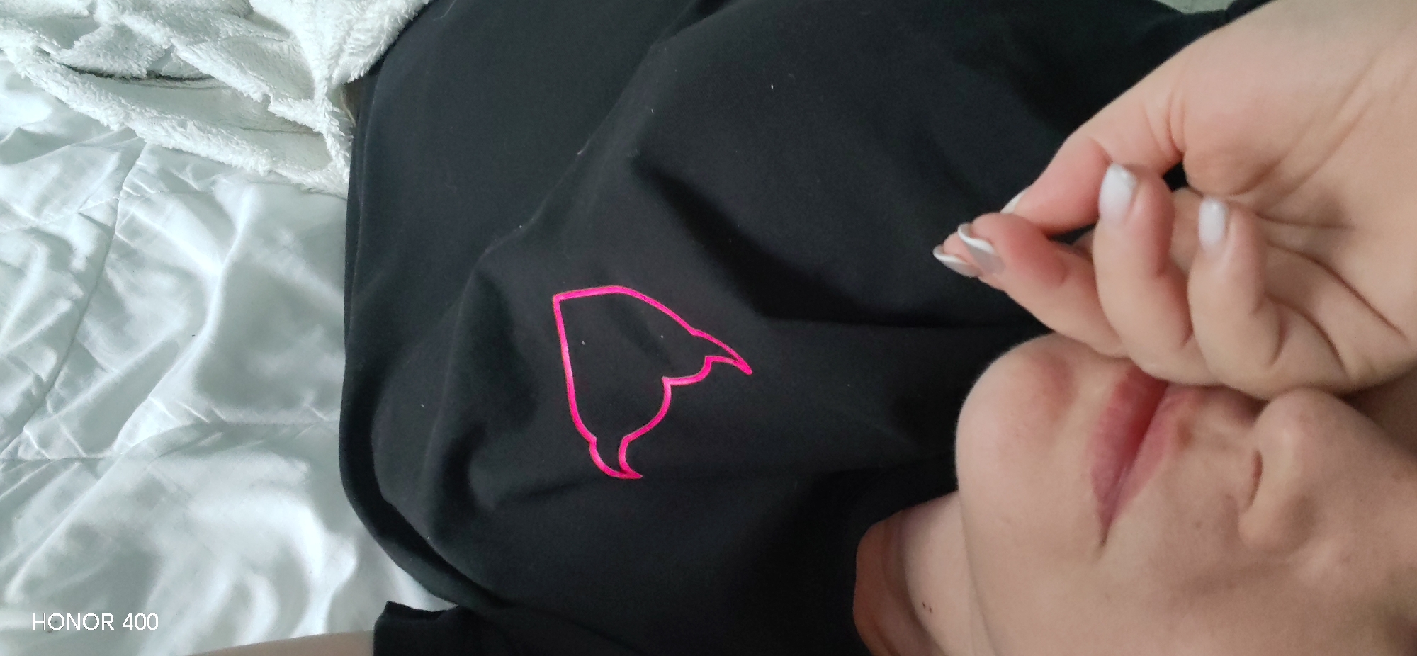 The image shows a person lying on a white bed. They are wearing a black garment with a pink design. Their hand is near their face. The scene does not reveal the city of Noviy, Russia.