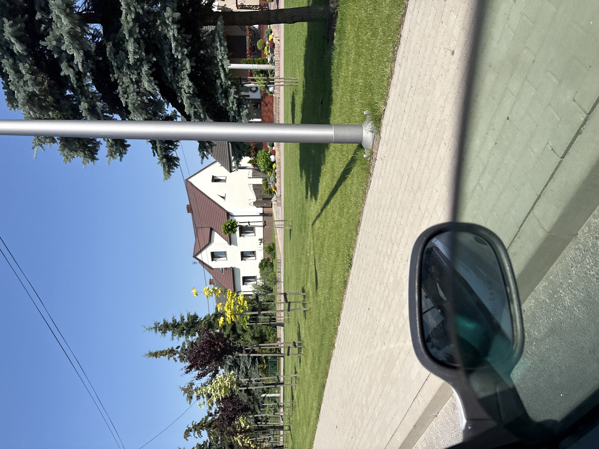 The image depicts a residential street scene in Mnichowice, Poland, on a clear, sunny day. In the foreground, a car's side mirror is visible on the left, reflecting the sky and the car's interior. Beyond the mirror, a paved sidewalk runs along a grassy area. A silver pole, likely a streetlight, stands prominently in the mid-ground, casting a shadow on the lawn. Several young trees, some with dark purple foliage and others with bright yellow leaves, are planted along the sidewalk, supported by stakes. In the background, two white houses with reddish-brown roofs are visible, surrounded by manicured lawns and decorative landscaping. The sky is a clear, vibrant blue, with two thin, dark power lines crossing the upper portion of the frame.