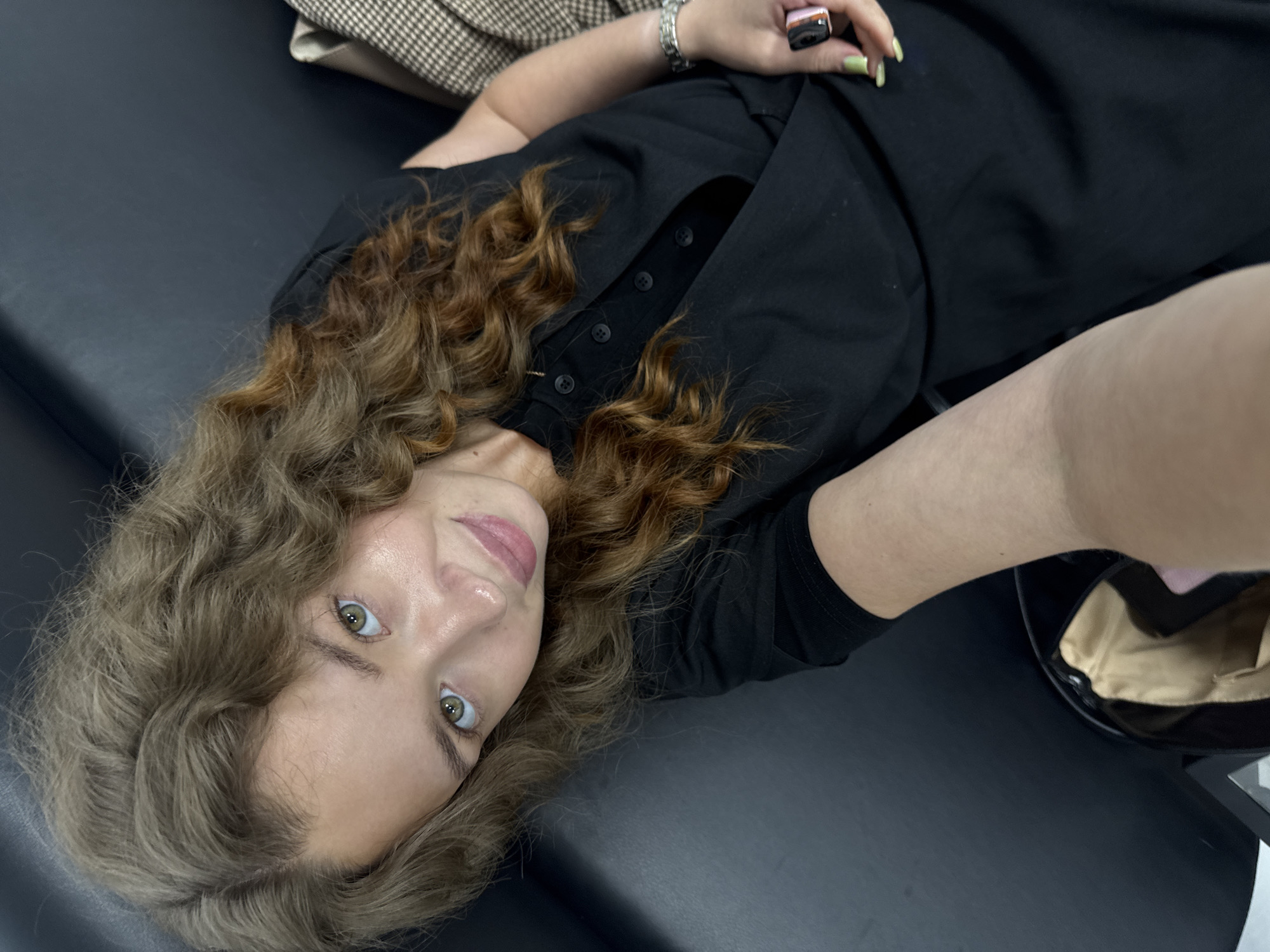 A young woman with curly brown hair and green eyes is pictured from a high angle, looking directly at the camera. She is wearing a black dress and is seated on a dark surface, possibly a couch or bench. A patterned fabric is visible behind her on the right side of the frame. Her right arm is extended forward, holding a dark object with pink and black accents, and her fingernails are painted light green. The background is dark and indistinct, suggesting an indoor setting. No specific location in the city of Chaikovsky, Russia can be determined from the image.