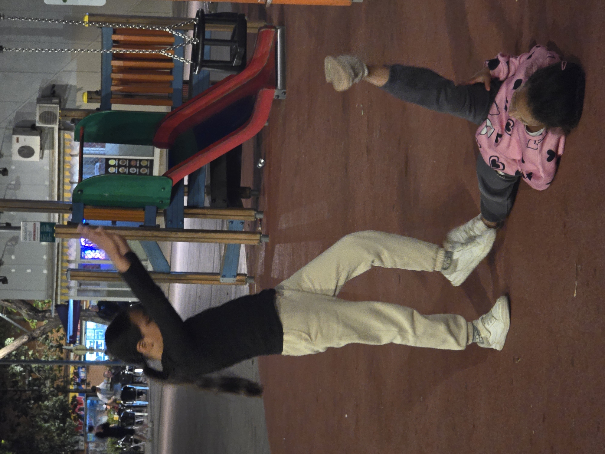 The image shows two children on what appears to be a playground in Cornellà De Llobregat, Spain. One child, wearing a black long-sleeved shirt and beige pants with white sneakers, is stretched out on the ground with their arms raised. Another child, wearing a pink heart-patterned sweatshirt and grey pants with beige sneakers, is also lying on the ground. The background includes playground equipment such as a slide and possibly other structures. The playground surface is brown, and there is a street scene visible in the background with trees and outdoor seating for restaurants or cafes.