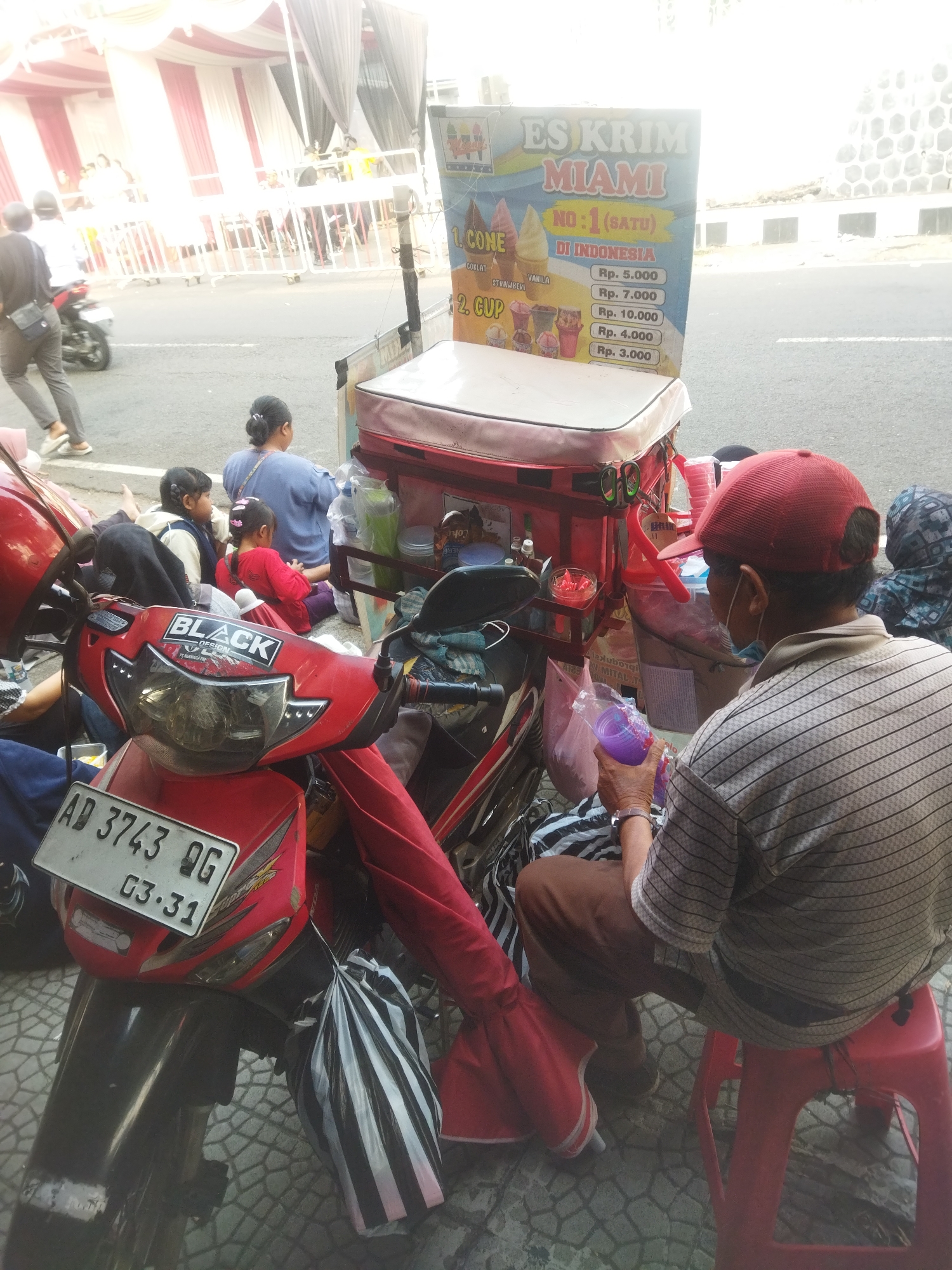 The image shows an outdoor scene on a street in Wonogiri, Indonesia, with an ice cream cart as the central subject. The cart is red and white and has a sign advertising "ES KRIM MIAMI". The sign lists prices for ice cream cones and cups. A man wearing a red cap and a striped shirt is seated on a red stool, holding a purple cup and what appears to be a plastic bag with ice cream. Behind him, several people are sitting on the ground, including a woman in a blue shirt and two young children, one in a red shirt. A red motorcycle with a license plate reading "AD 3743 OG" is parked in front of the ice cream cart. The background shows a street, a sidewalk, and buildings, with some people and a motorcycle in the distance. The overall impression is of a typical street scene with a vendor and customers in a tropical environment.