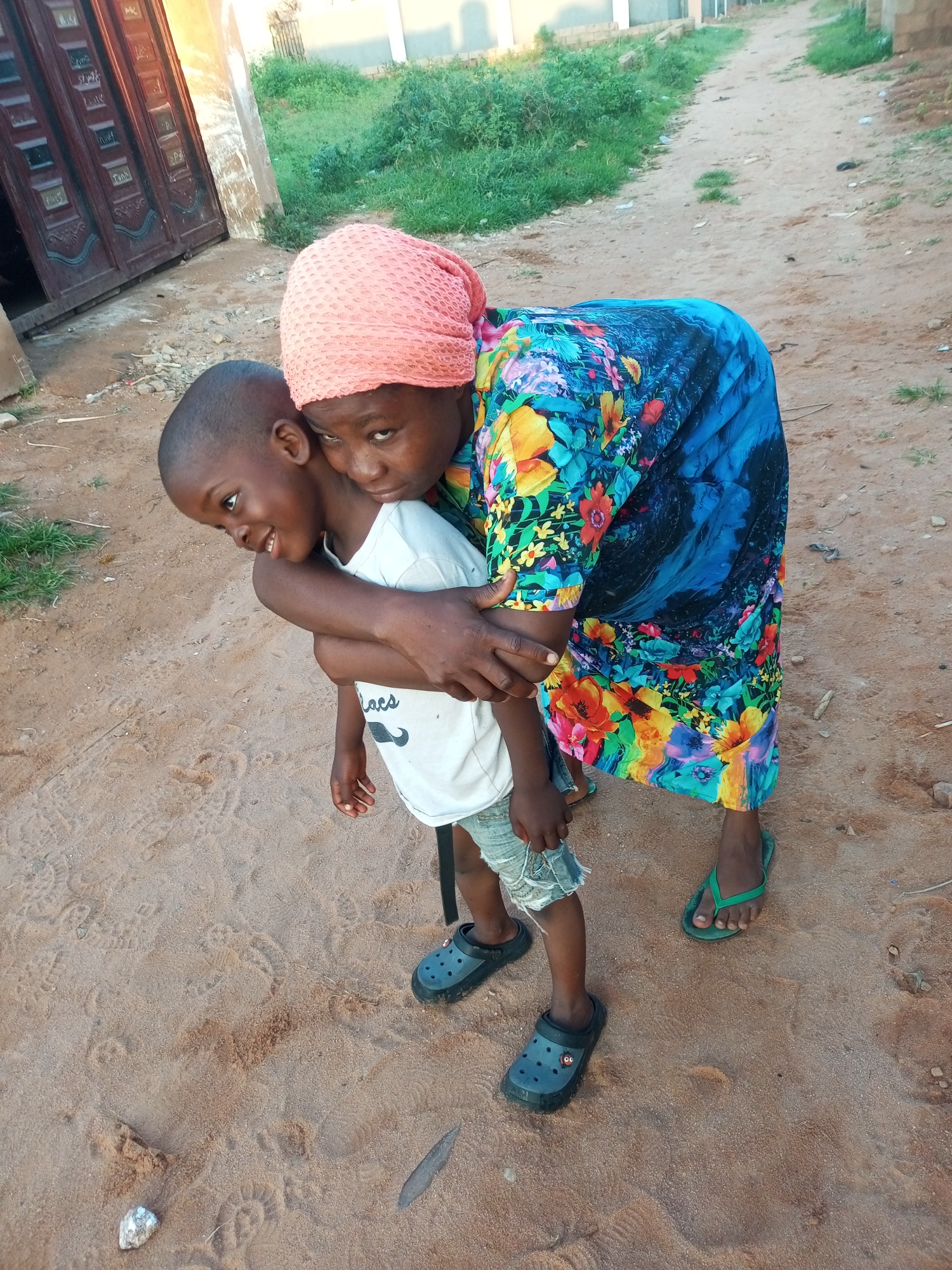 A woman in a pink headscarf and a vibrant, floral-patterned dress is giving a young boy a piggyback ride. She is embracing him from behind as he looks to his left with a smile. He is wearing a white t-shirt and distressed denim shorts, and both are wearing dark-colored clogs. The woman has green flip-flops on her right foot. They are standing on a sandy ground with some footprints and small stones. In the background, there is a building with dark wooden doors, green vegetation, and a dirt path. The scene appears to be outdoors, possibly in a residential area, and the lighting suggests it is daytime. The overall mood seems affectionate and joyful. There is no visible text in the image.