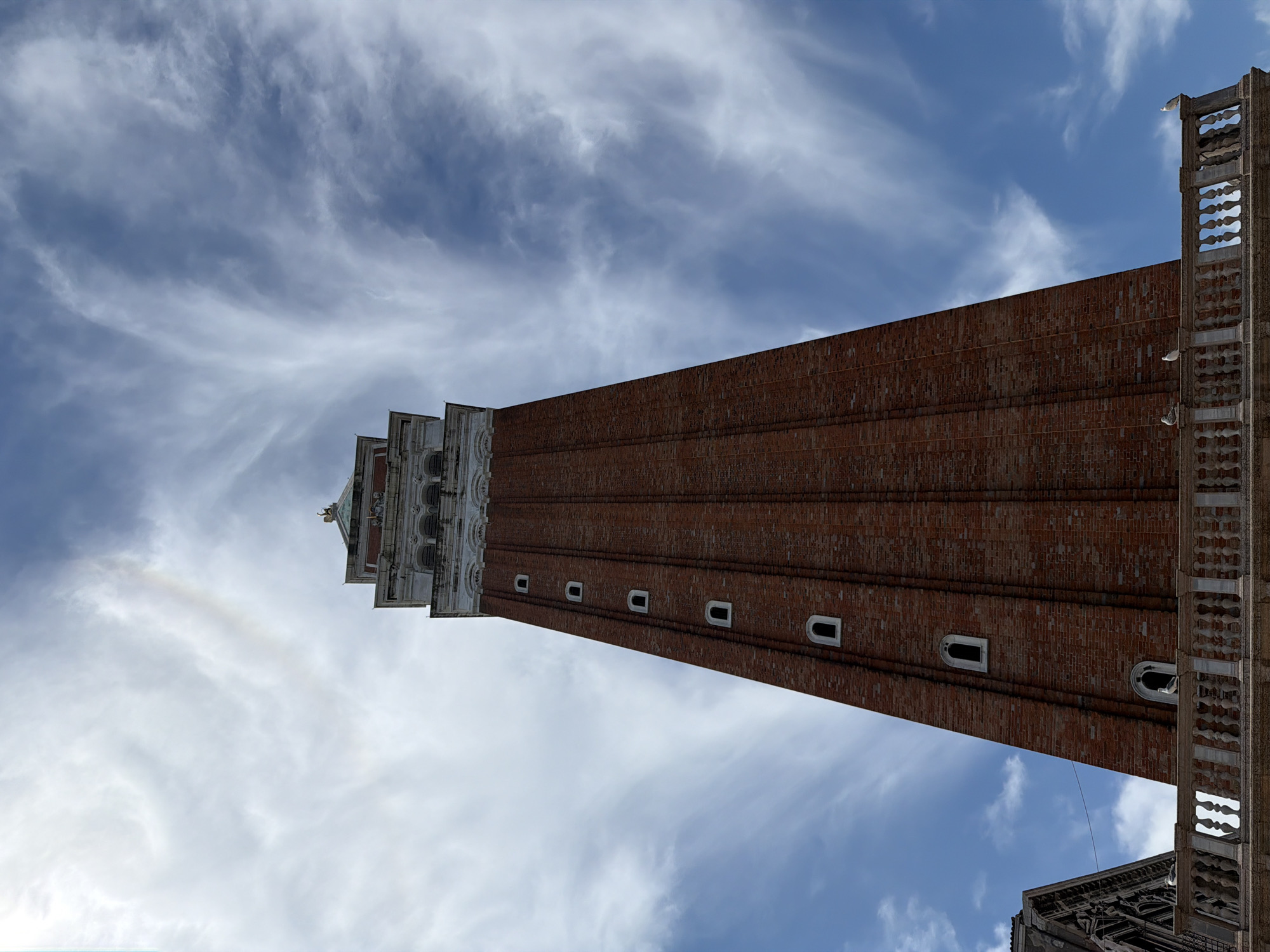 This is a low-angle, full shot of the St. Mark's Campanile in Venice, Italy, on a partly cloudy day. The campanile is a tall, red brick tower with white decorative accents at the top. It has numerous small, arched windows evenly spaced along its height. The top of the tower features a white, ornate structure with arches and decorative elements, topped by a pyramidal spire with a golden angel statue. A faint rainbow or atmospheric optical phenomenon is visible in the sky above and slightly to the left of the tower's apex. The sky itself is a vibrant blue with wispy, white clouds that create a dramatic effect. In the foreground, a white stone balustrade with decorative pillars runs horizontally across the bottom of the frame, suggesting a viewpoint from a balcony or elevated platform. Three seagulls are perched on the balustrade. To the far left, a portion of a gothic-style building with intricate stone carvings is visible. The image captures the grandeur of the campanile against a dynamic sky.