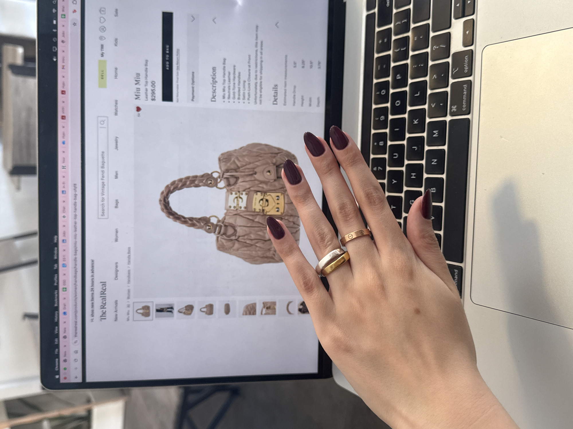An overhead shot captures a person's left hand interacting with a silver Apple MacBook in what appears to be an indoor setting, likely a desk. The hand, adorned with dark red, almond-shaped nail polish, wears two rings on the ring finger: a slim gold band (resembling a Cartier Love Ring) and a wider two-tone ring with gold and silver sections.
The laptop screen displays the website "The RealReal," an online luxury consignment store. The active page features a "Miu Miu Leather Top Handle Bag" priced at "$295.00," with a detailed description including "Neutrals Leather," "Gold-Tone Hardware," "Braided Handles," "Satin Lining," and "Push-Lock Closure at front." The description also notes, "Unfortunately, due to restrictions, this item may not be eligible for shipping in all areas." Estimated item measurements for "Handle Drop," "Height," "Width," and "Depth" are visible (6.5", 8.5", 10.0", 3.0" respectively). The website's navigation includes categories like "Watches," "Jewelry," "Men," "Bags," "Women," "Home," "Kids," "Sell," and "My TRR." A search bar at the top shows "Search for Vintage Fendi Baguette." Various browser tabs are open, including "the Hype." The right side of the image shows part of the laptop's QWERTY keyboard with keys such as "command" and "option."
The scene is set in Mexico City, Mexico.
