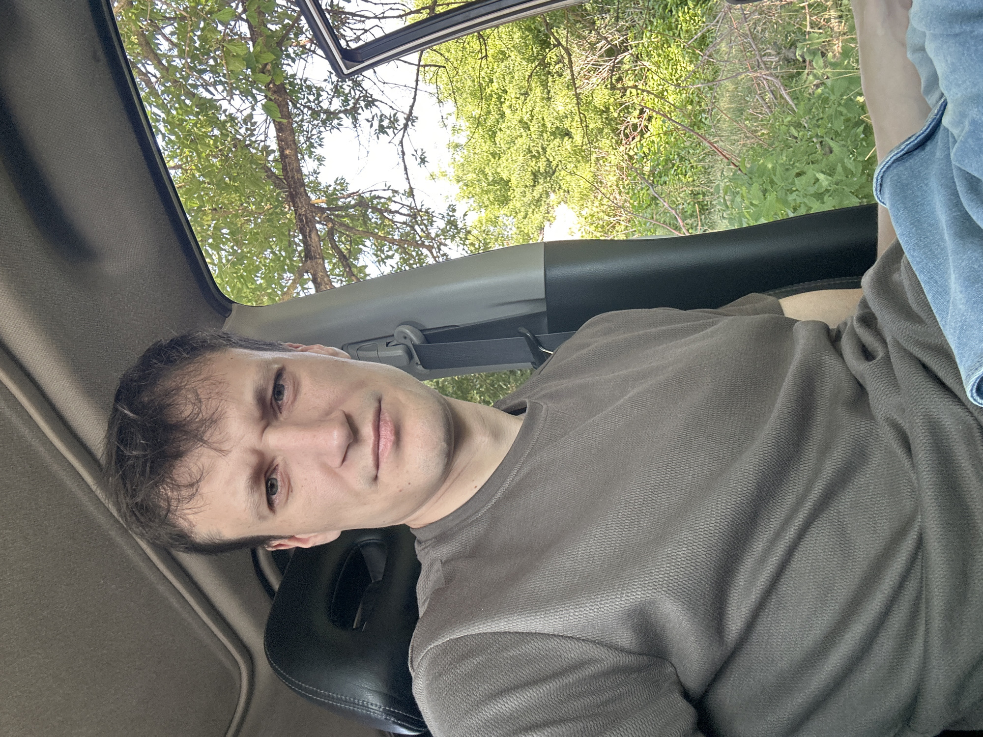 A man is sitting in the driver's seat of a car, looking directly at the camera. He is wearing a brown t-shirt and blue jeans. The car appears to be parked on the side of a road, with trees and foliage visible through the open passenger side door and the windshield. The sky is clear and bright. The image was taken outdoors in Makiv, Ukraine.
