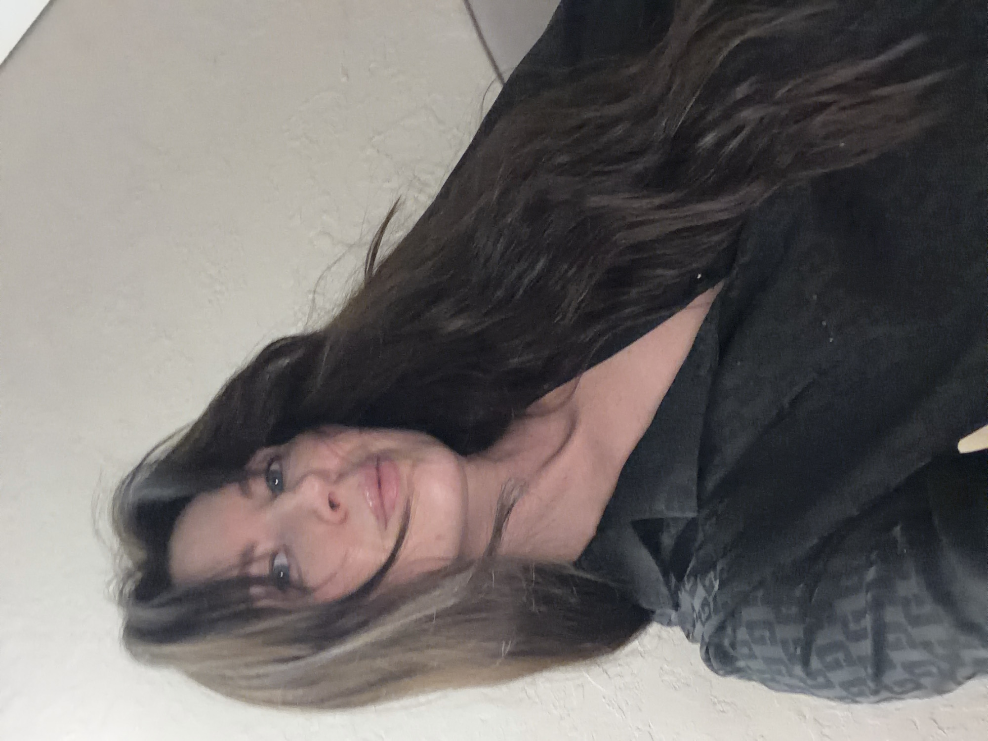 A woman with long, dark and light brown hair is lying on her side, facing the camera. She is wearing a dark-colored garment. The background is a light-colored wall. The location in the city of Perm, Russia, is not discernible from the image.