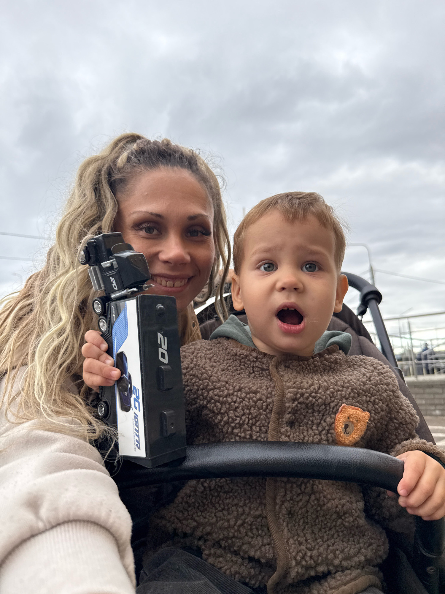 In Bor, Russia, a woman with blonde hair in a ponytail is holding a black toy truck with white and blue detailing. She is smiling and looking towards the camera. Beside her, a young child with blonde hair is seated in a stroller, wearing a brown, fuzzy jacket with a teddy bear patch. The child's mouth is open in surprise or excitement. The background appears to be an outdoor, overcast environment, possibly a park or street, with a railing and blurred figures in the distance. The overall impression is a candid, affectionate moment captured between a mother and her child.