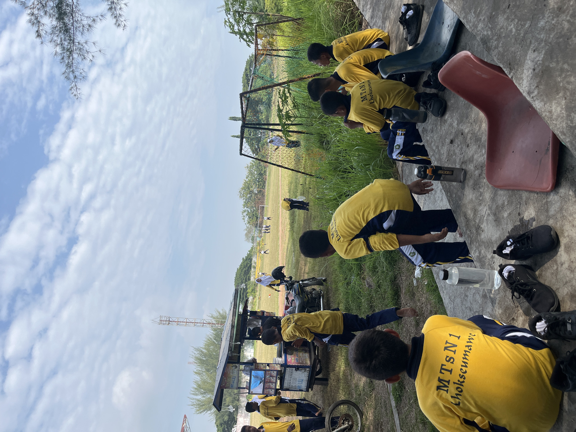 In Lhokseumawe, Indonesia, a group of students in yellow and navy blue uniforms are gathered on a grassy field, likely part of a school or sports complex. Several students are seated on a concrete ledge in the foreground, facing away from the camera. Their shirts bear the inscription "MTsN 1 Lhokseumawe," identifying their school. Scattered around them are athletic shoes and water bottles. In the mid-ground, a food cart is set up on a paved area, with students interacting around it. Further into the field, other students are engaged in activities, with some playing and others observing. The environment appears to be outdoors, with a clear sky, scattered clouds, and trees in the background. A sports goal structure is visible on the right side of the frame. The overall scene suggests a casual, possibly break time, during a school event or sports practice.