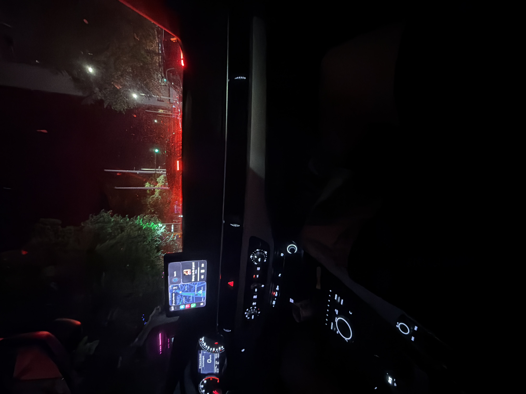 The image displays the interior perspective from a stationary vehicle at night, looking forward through the windshield onto a city street in Ruse, Bulgaria, at approximately 20:17, as indicated on the car's infotainment screen.
**Vehicle Interior:** The dashboard of a modern car is visible, featuring an illuminated digital instrument cluster displaying "0 km/h," indicating the vehicle is stopped. A central infotainment screen shows a navigation map application with a blue route, alongside what appears to be a music player displaying "Surey yo de el parvi." Below the screen, illuminated climate control dials, buttons, and a central rotary controller are visible. A red hazard light button is present but unlit. Ambient lighting, possibly a strip of white LEDs, is subtly visible on the passenger side of the dashboard. Portions of a steering wheel are visible on the left. The lower right portion of the frame shows a dark, indistinct form, possibly a passenger's leg or arm.
**Exterior Scene:** The view through the windshield reveals a dark urban environment illuminated by artificial lights. Multiple vehicles are stopped ahead, their red taillights distinctly visible across the width of the road. A prominent green light, consistent with a traffic signal, is visible on the left side of the frame, above the line of stopped cars. Streetlights illuminate portions of the surrounding trees, which show green foliage. Some foliage also reflects a distinct green glow, possibly from another light source. The windshield appears to have minor streaks or reflections, but no heavy precipitation is evident. The overall scene depicts a traffic queue in a city at night.