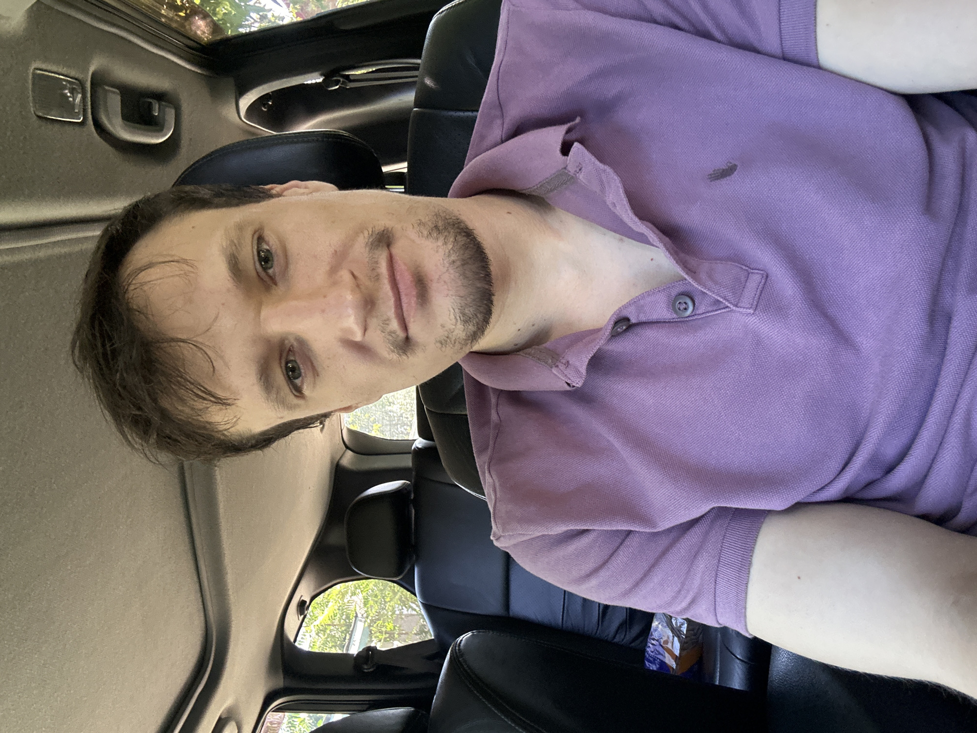 A man with short brown hair, stubble, and green eyes is seated in the passenger seat of a car. He is wearing a purple polo shirt. The interior of the car is visible, including black leather seats and the roof. Sunlight filters in from the windows, illuminating trees and foliage outside. The image appears to be taken from a selfie perspective. The location is Makiv, Ukraine.