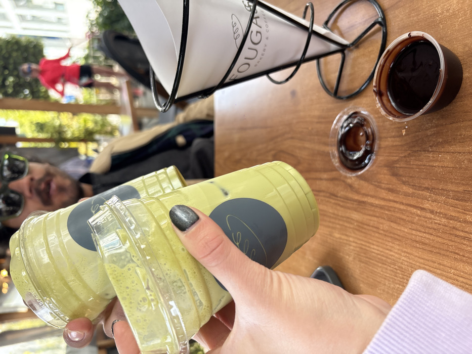 Two clear plastic cups filled with a green beverage are held by a hand with glittery gray nail polish. On the wooden table, a metal cone-shaped holder contains paper cones labeled "Fouga Bakery & Coffee". Nearby are two small containers of dark liquid, likely chocolate sauce. A man with sunglasses lies on a bench in the background, while another person in red stands on a walkway above.