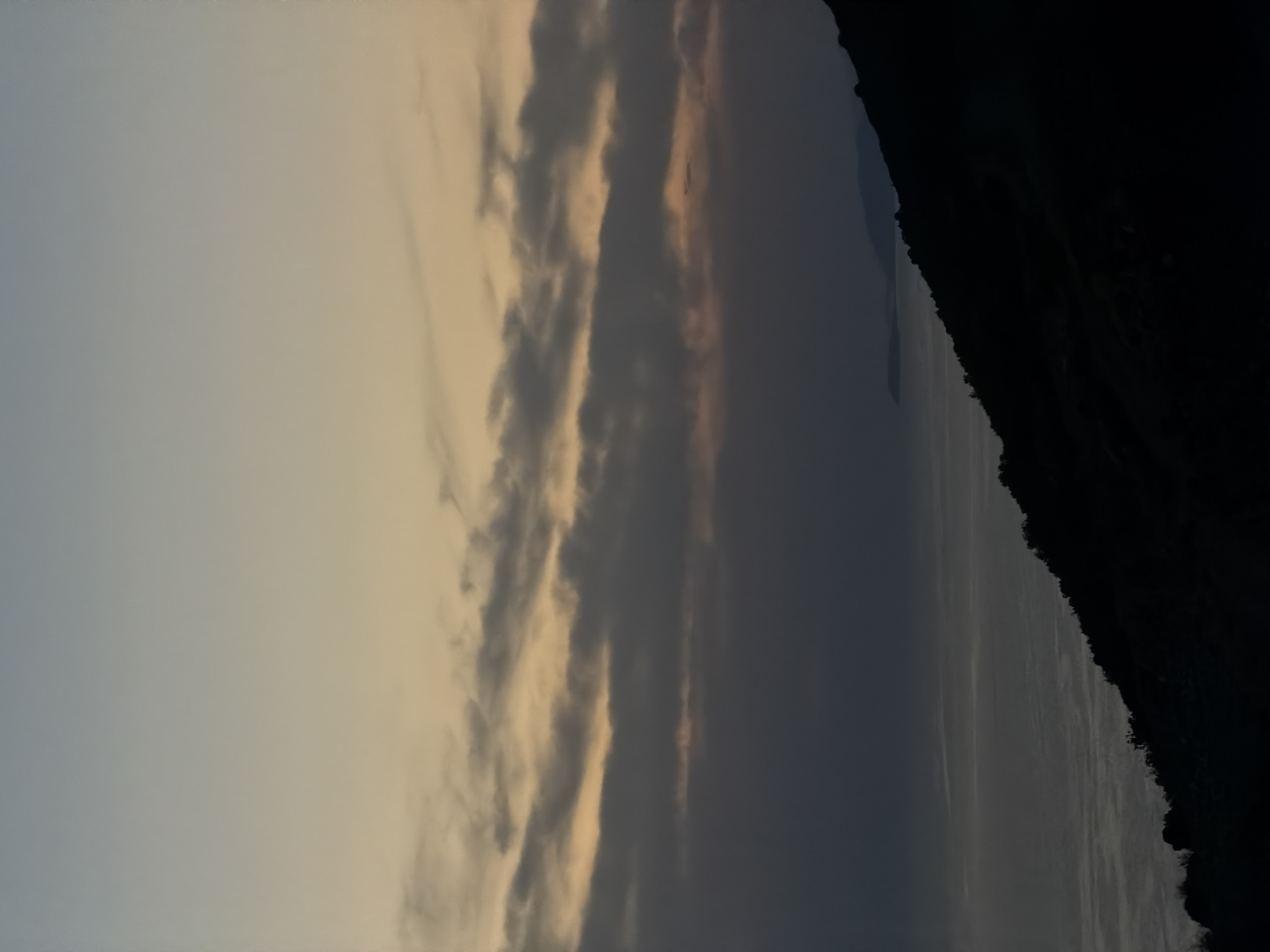 The image depicts a twilight scene over the sea, with a silhouetted mountain range in the foreground. The sky transitions from a pale blue at the top to a muted orange and yellow near the horizon, with streaks of dark clouds scattered across it. Below the clouds, a layer of mist or fog obscures the lower portion of the sky and merges with the distant sea. The sea itself is a dark, choppy expanse, reflecting the dim light from the sky. The mountain on the right side of the frame is a stark silhouette, with its rugged outline sharply defined against the lighter sky and sea. Its lower slopes are covered in what appears to be sparse vegetation. In the distance, across the water, other mountain formations are faintly visible, also rendered in shadow. The overall impression is one of serene, natural beauty at the end of the day. The location specified, Zagora, Montenegro, suggests a coastal mountain village, which aligns with the visual elements of the image.