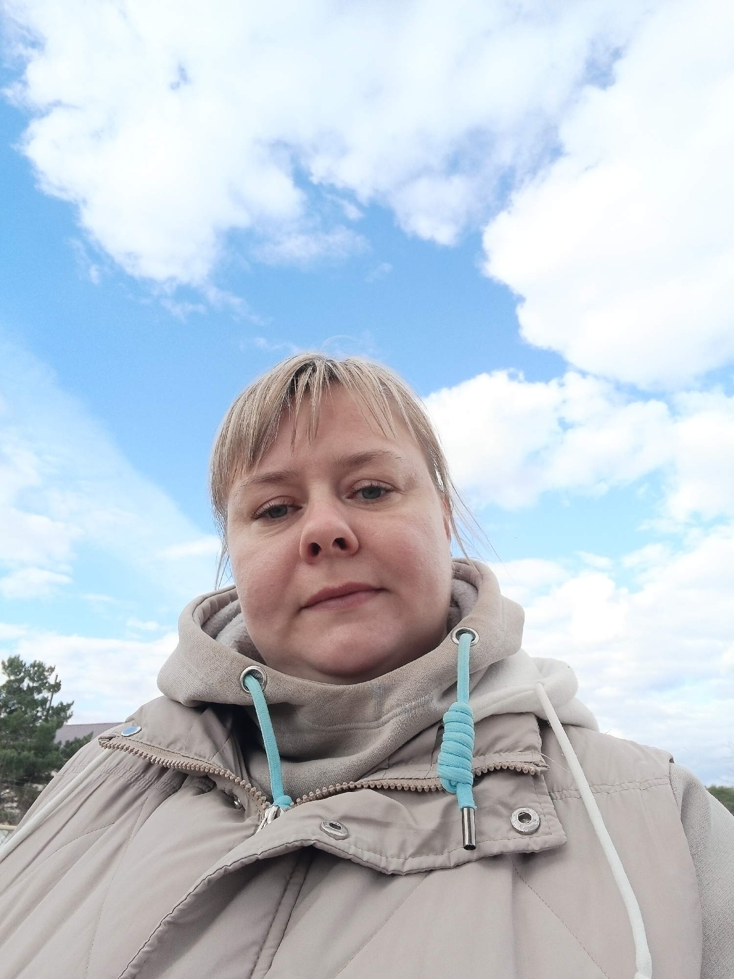 A close-up selfie captures a woman from the chest up, looking directly at the camera with a neutral expression. She has light-colored hair with bangs. She is wearing a light beige or tan quilted vest over a matching hooded sweatshirt. A notable detail of her clothing is the light blue drawstring of the hoodie, which is tied in a knot and has silver-toned metal tips. The vest features a zipper and visible snap buttons.
The background is dominated by a bright blue sky filled with scattered white, fluffy clouds, indicating daytime with fair weather. In the lower-left corner, a small glimpse of green foliage, likely from a tree, and a distant building are visible, suggesting an outdoor, possibly urban or suburban, setting in Chaikovsky, Russia. No discernible text is visible in the image.