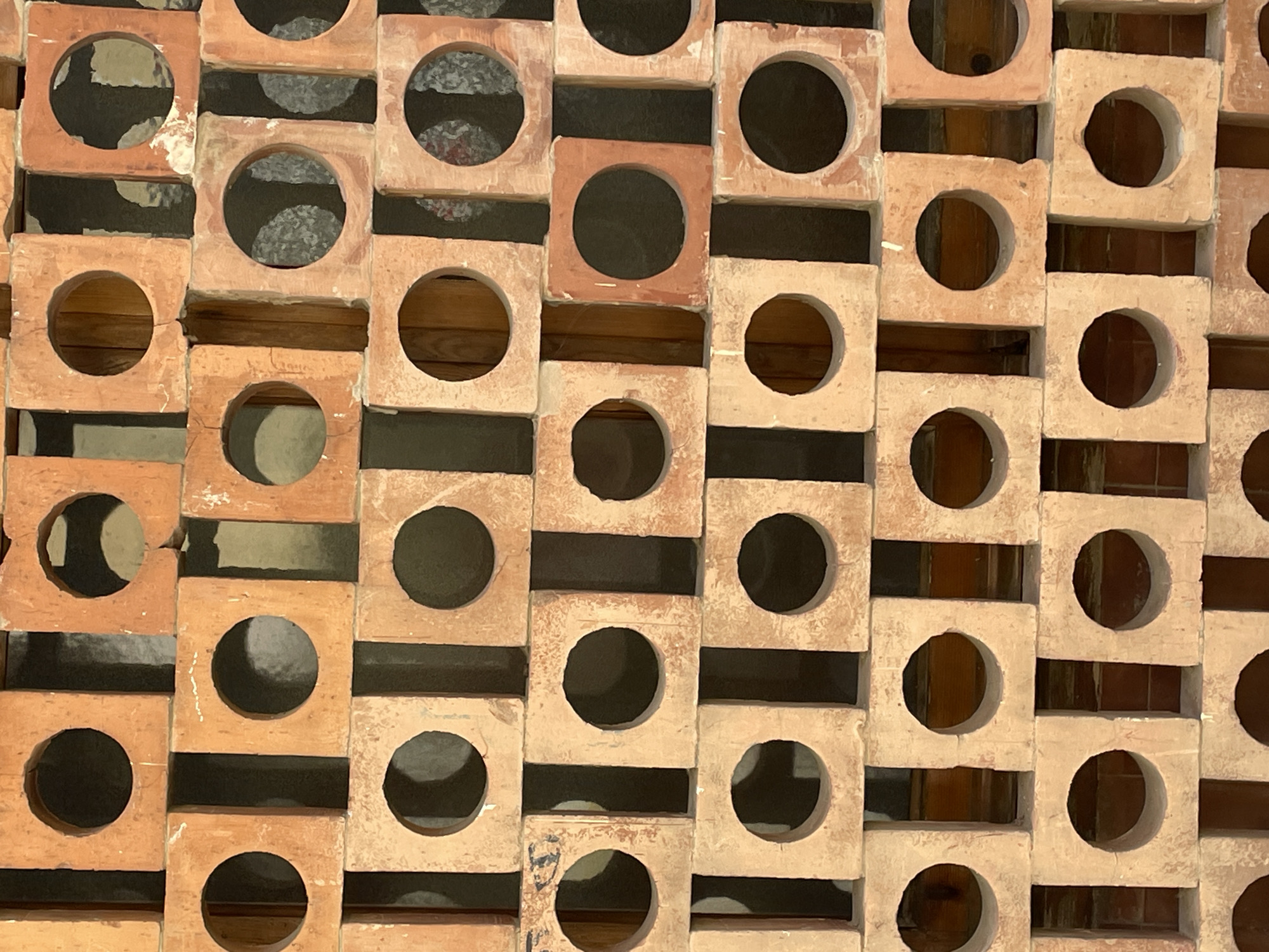 This media file shows a close-up of a decorative wall constructed from square, terracotta-colored bricks, each with a circular hole in the center. The bricks are arranged in a repeating pattern, with alternating rows offset from each other. This creates an open, geometric design, allowing light and shadow to play across the surface. The lighting suggests it is daytime, possibly with diffused sunlight, as there are no harsh shadows. The overall impression is one of textured, artisanal construction. The material and style are reminiscent of traditional Spanish architecture, potentially found in a location like Barcelona, Spain, where such decorative screens are common in courtyards or as architectural features. No people or text are visible in the image.