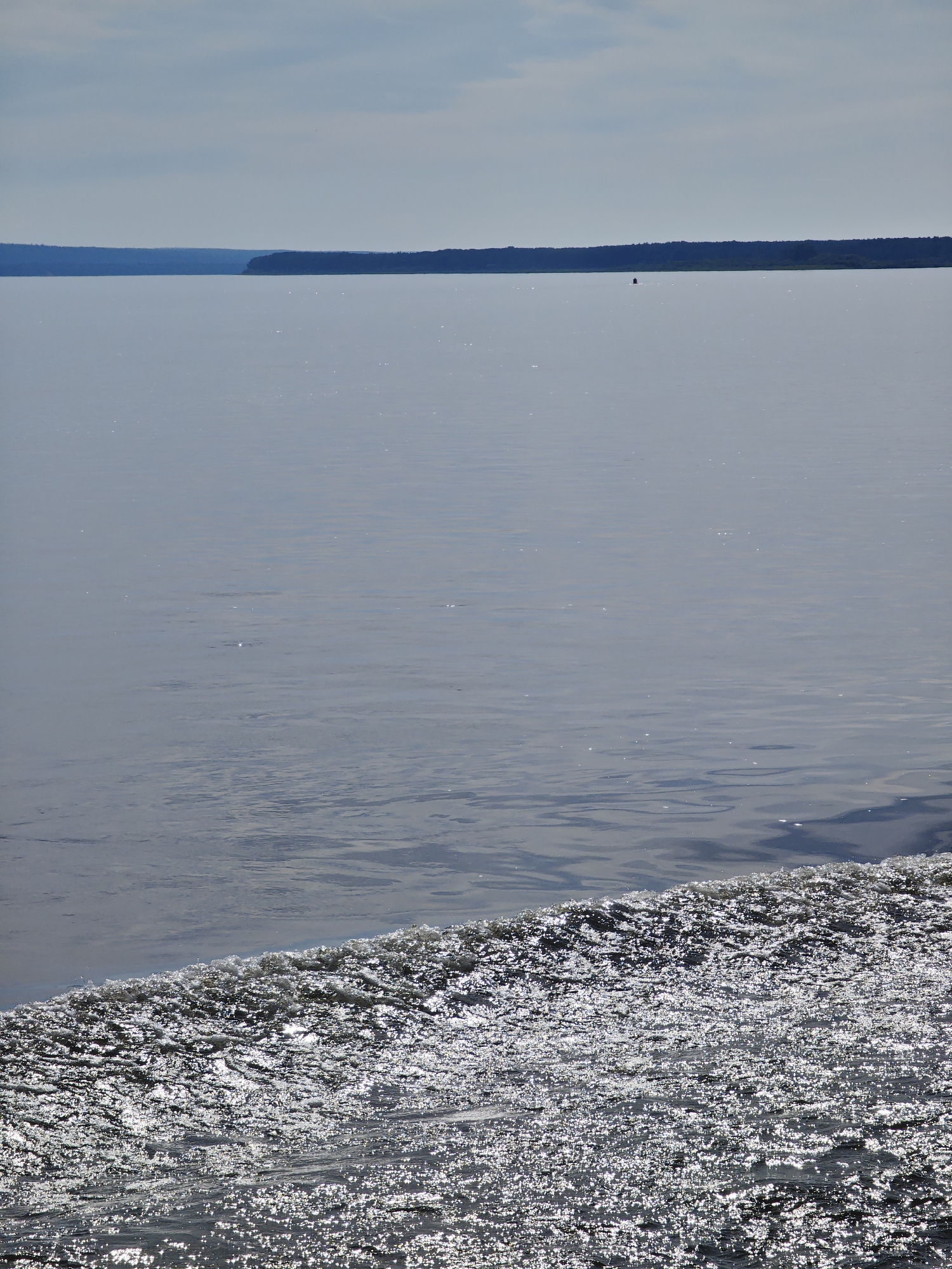The image shows a wide body of water, likely a river or lake, with a distant, tree-lined shore. The water in the foreground is choppy and reflects the sunlight, creating a shimmering effect. In the background, a dark, horizontal line represents the distant landmass. The sky above is partly cloudy with a hazy, diffused light. There is a small, dark marker or buoy visible on the water in the far distance. The scene is tranquil and natural, suggesting an outdoor environment. No specific landmarks or structures are identifiable that would definitively place it in Yartsevo, Russia. The image captures a moment of calm on the water, with the dynamic play of light on the surface providing visual interest.