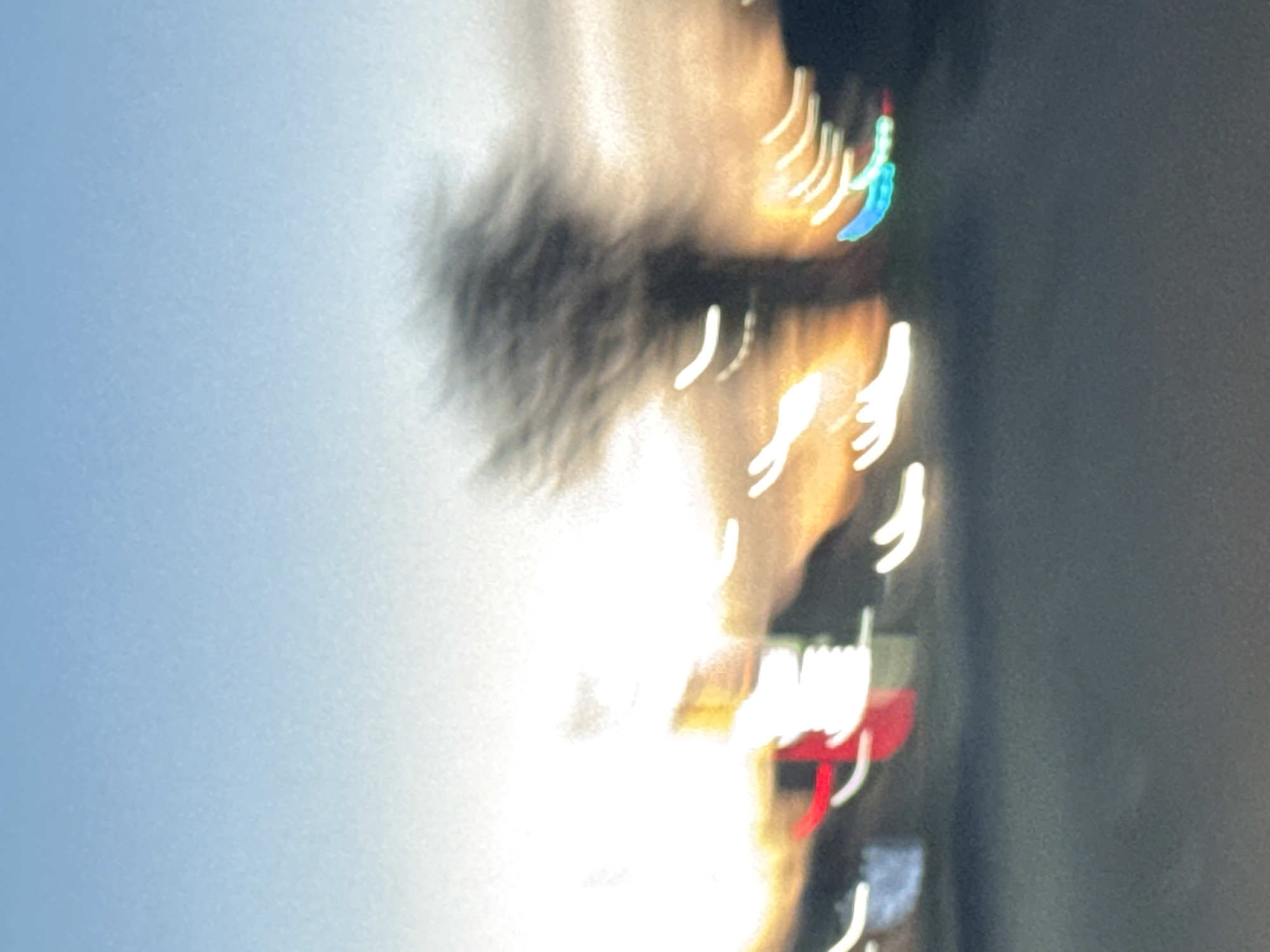 The image appears to be a blurry, dynamic shot taken while in motion, possibly from a moving vehicle. On the left, there's a pale blue sky. In the central area, there's a hazy figure that looks like a person with dark, possibly long, hair. This figure is blurred, suggesting movement. To the right, there are streaks of light, some white and some with hints of other colors like red and blue. These streaks likely represent car lights or city lights. The background on the right is a dark gray color, possibly representing the side of a building or a dark road. The location in Chaikovsky, Russia is unclear based on this image alone. More context or specific details visible in a clearer image would be needed to identify the specific location.