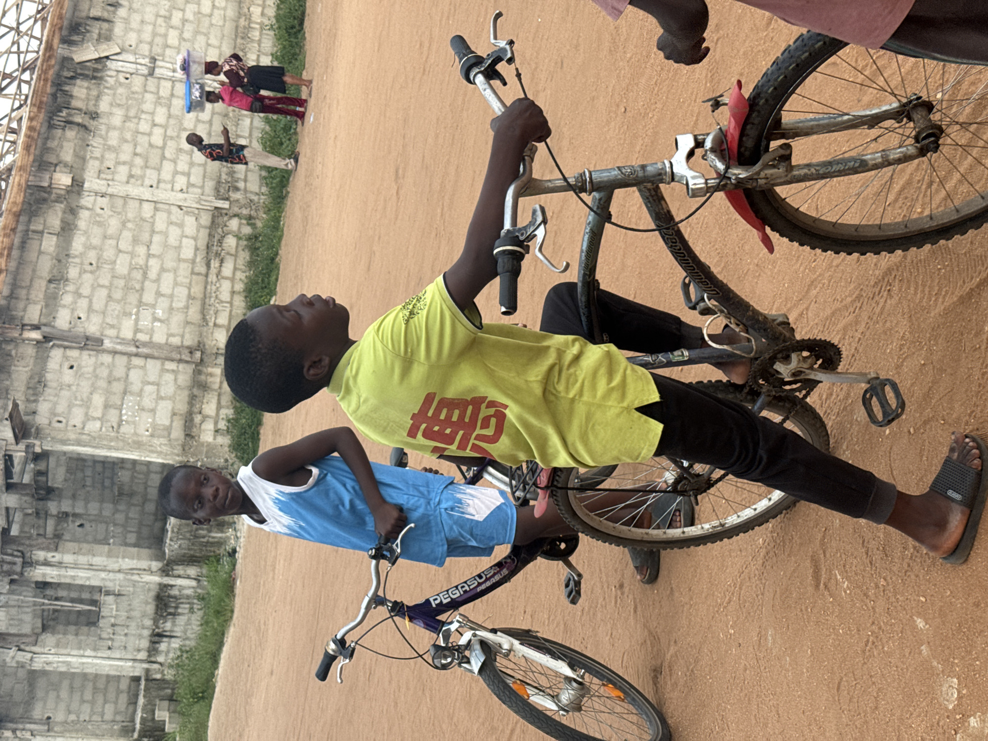 This outdoor photograph, taken in Ihiagwa, Nigeria, features two young boys with bicycles in the foreground against a backdrop of a sandy, unpaved area and a concrete block wall. The image is rotated, showing the subjects and scene vertically.
In the immediate foreground, a boy wearing a bright yellow t-shirt with red, possibly East Asian, characters printed on it stands holding the handlebars of a dark-framed bicycle with a red mudguard. He wears black pants and black flip-flops. His gaze is directed to the left, away from the camera. Beside him, another boy in a blue and white tank top holds the handlebars of a blue bicycle with "PEGASUS" written on its frame. This boy looks directly at the camera with a neutral expression. Both bicycles appear to be utility or mountain bike styles.
The setting is characterized by a dusty, reddish-brown ground, indicative of a dry climate. In the background, a partly constructed or older building made of concrete blocks is visible, with some sparse green vegetation growing at its base. Several adults are present in the mid-ground: one person in a patterned shirt and light pants, and two women, one of whom is carrying a container or bucket on her head. Another figure in a pink/red top is also visible. The people in the background appear to be walking or standing casually.
The lighting suggests it is daytime, with diffused light indicating either an overcast sky or an early/late sun. The overall scene depicts a typical day in a community setting, with children and adults going about their activities.