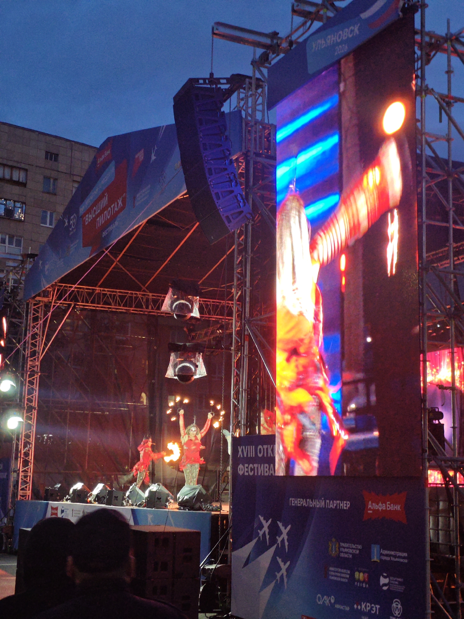 This image depicts an outdoor event, likely a festival, at dusk or evening in Ulyanovsk, Russia, as indicated by the text "УЛЬЯНОВСК 2026" on a large LED screen. A stage is set up with performers on it, one of whom is in a red costume and appears to be holding a flaming prop. Another performer, also in red, is to their right. There are large speakers suspended from scaffolding above the stage. The main LED screen displays a vibrant, abstract image with predominantly blue and red tones, and what appears to be a blurry depiction of a person in costume. Below the LED screen is a banner with logos and text, including "ГЕНЕРАЛЬНЫЙ ПАРТНЕР" (General Partner) and "Альфа Банк" (Alfa-Bank). Several individuals are visible in the foreground, their backs turned to the camera, suggesting they are part of the audience. The overall atmosphere appears to be festive and energetic, with the stage lighting and the bright LED screen contributing to the visual spectacle.