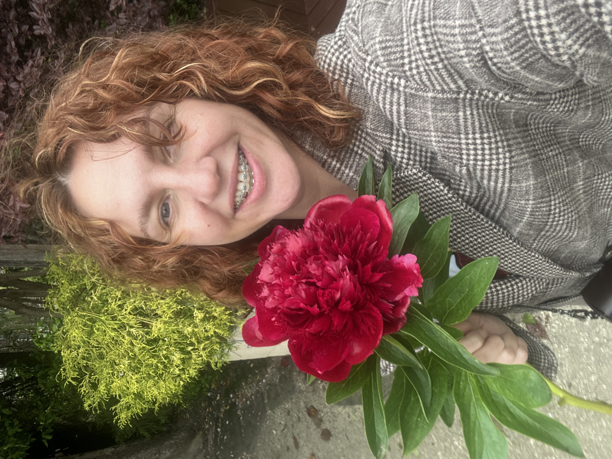 A woman with curly auburn hair and braces smiles while holding a large red peony. She is wearing a gray plaid jacket. The background includes greenery, possibly bushes and trees, and a paved surface. The location cannot be confirmed as Sochi, Russia.