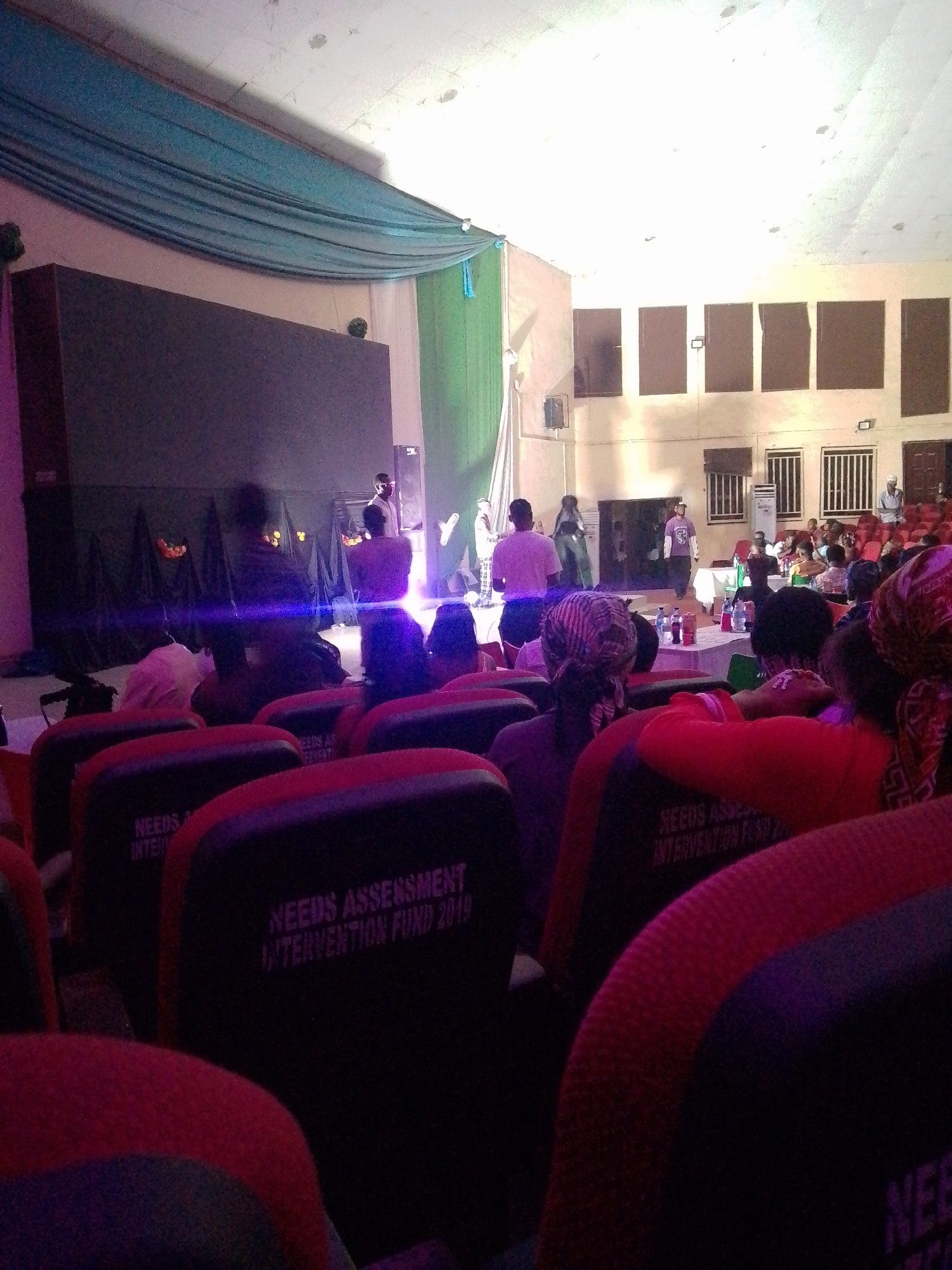 The image shows an indoor auditorium with a stage and an audience seated in red and black chairs. On the stage, several people are present, some appearing to be performers or speakers. There is a large dark screen at the back of the stage, and decorative draping in shades of blue and green above it. The lighting suggests an evening event. The audience members are partially visible, with many facing the stage.
Some of the seats in the foreground have text printed on them, reading "NEEDS ASSESSMENT INTERVENTION FUND 2019". The overall atmosphere appears to be that of a formal gathering, presentation, or performance. The lighting is somewhat dramatic, with purple and blue hues visible on the stage and in the foreground. The people in the audience are mostly focused on the activity on stage, and their expressions are not clearly discernible. The setting is likely a conference hall, theater, or similar venue.