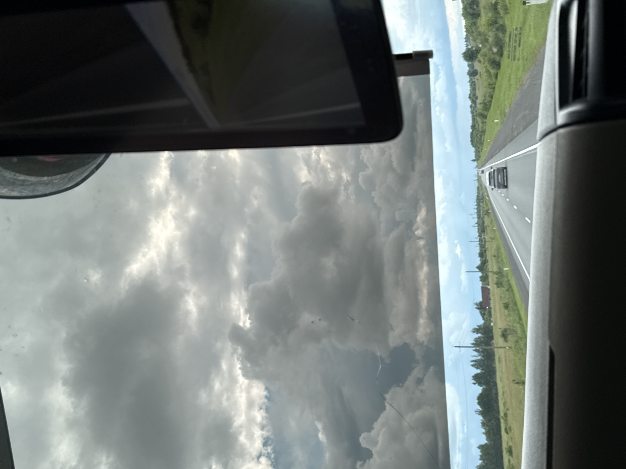 The image shows a view from inside a vehicle driving on a highway. The sky is overcast with dramatic dark clouds. The highway stretches into the distance, with a few other vehicles visible. The sides of the road are lined with green fields and trees, and some power lines and buildings are scattered throughout the landscape. A dark screen, possibly a GPS or entertainment system, is visible on the right side of the frame, partially obscuring the view. The location is identified as Kipuya, Russia.