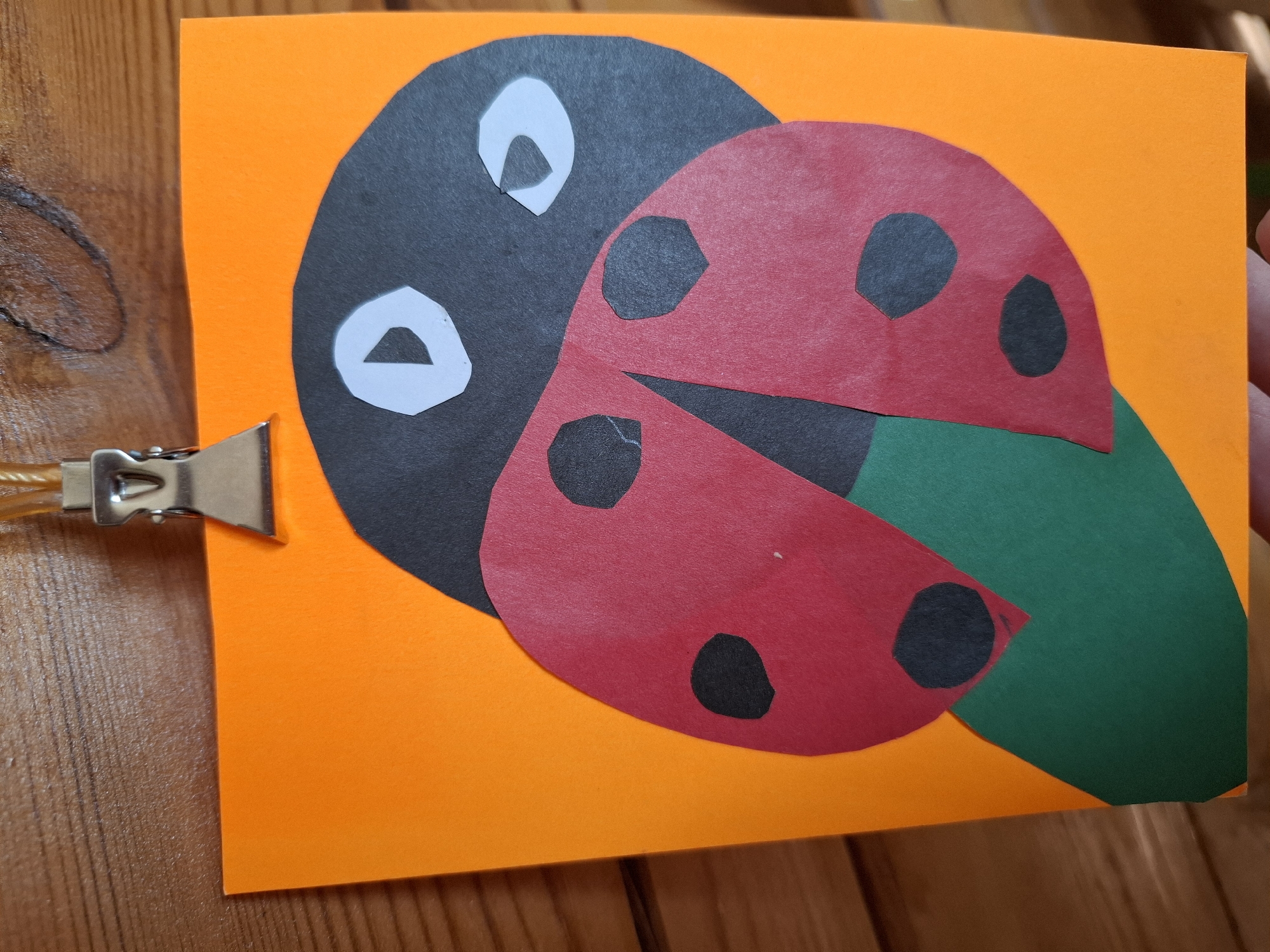 A handmade craft project, likely a ladybug, is displayed on an orange paper background. The ladybug is constructed from black, red, and green construction paper, with two white paper circles representing eyes. The project is attached to a wooden surface with a silver clip. The image does not contain information about the city of Tyumen, Russia.