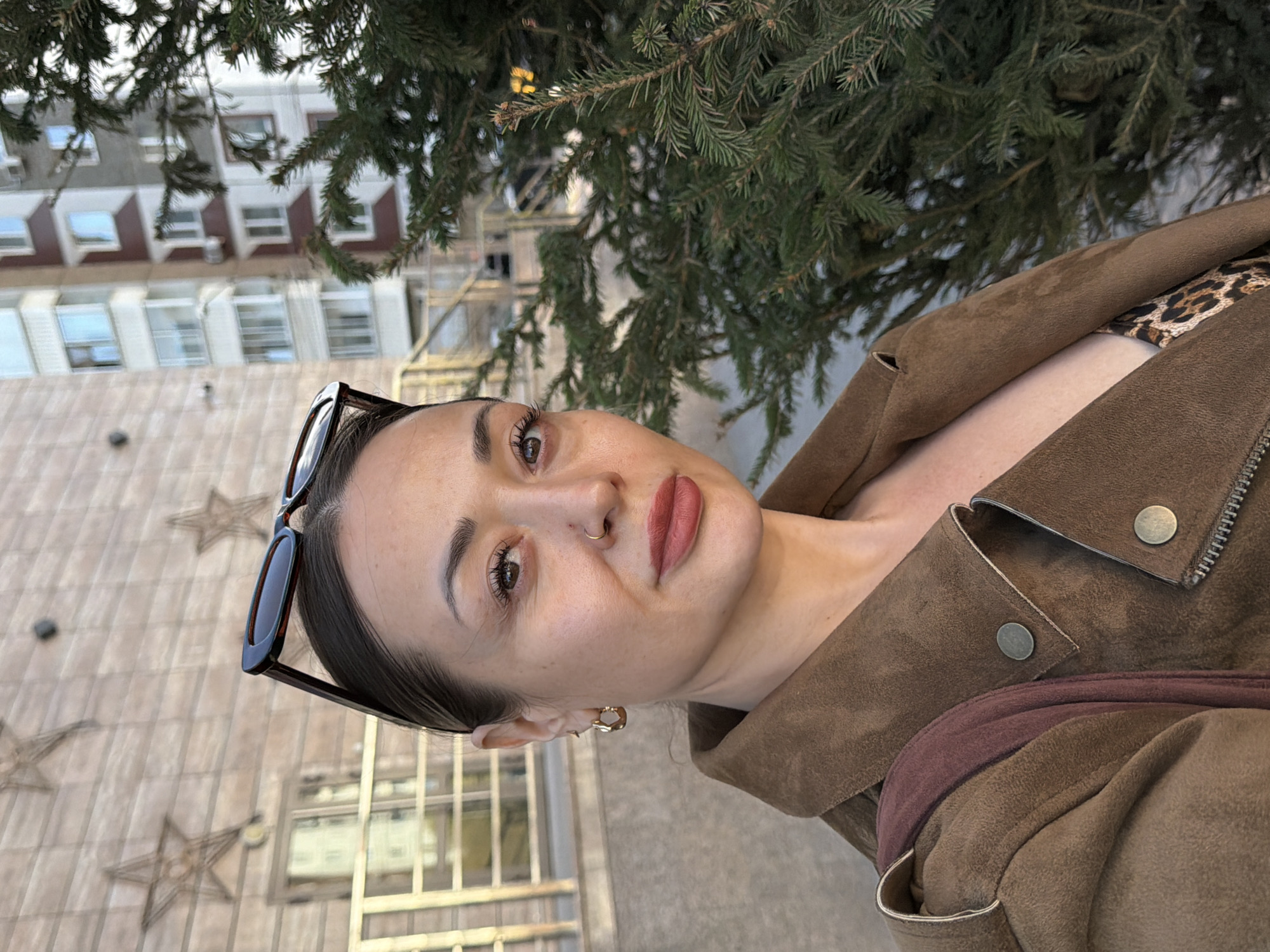 A woman is taking a selfie outdoors. She is wearing a brown suede jacket over what appears to be a leopard print top. Sunglasses are perched on her head. The background includes a building decorated with stars, a fir tree, and other buildings. It's daytime, and the location is identifiable as Krasnoyarsk, Russia, given the distinctive building architecture and decorative elements.