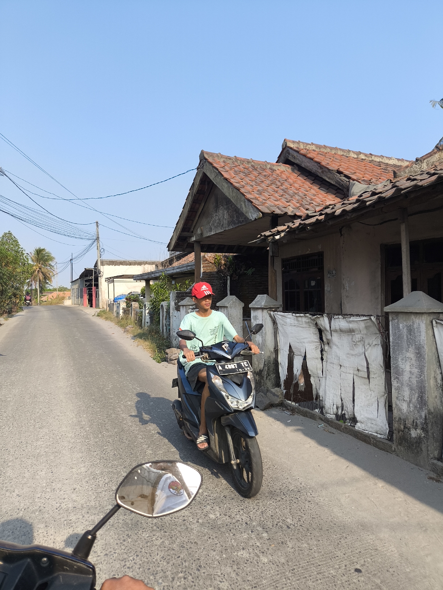 A young male wearing a red cap and a light green t-shirt rides a dark blue scooter down a paved road lined with houses. The scooter has a license plate that reads "A 4897 TC". The houses have red tile roofs and weathered walls. The road is flanked by fences and vegetation. The sky is clear and blue. The photo appears to have been taken in a residential area of Cilegon, Indonesia.