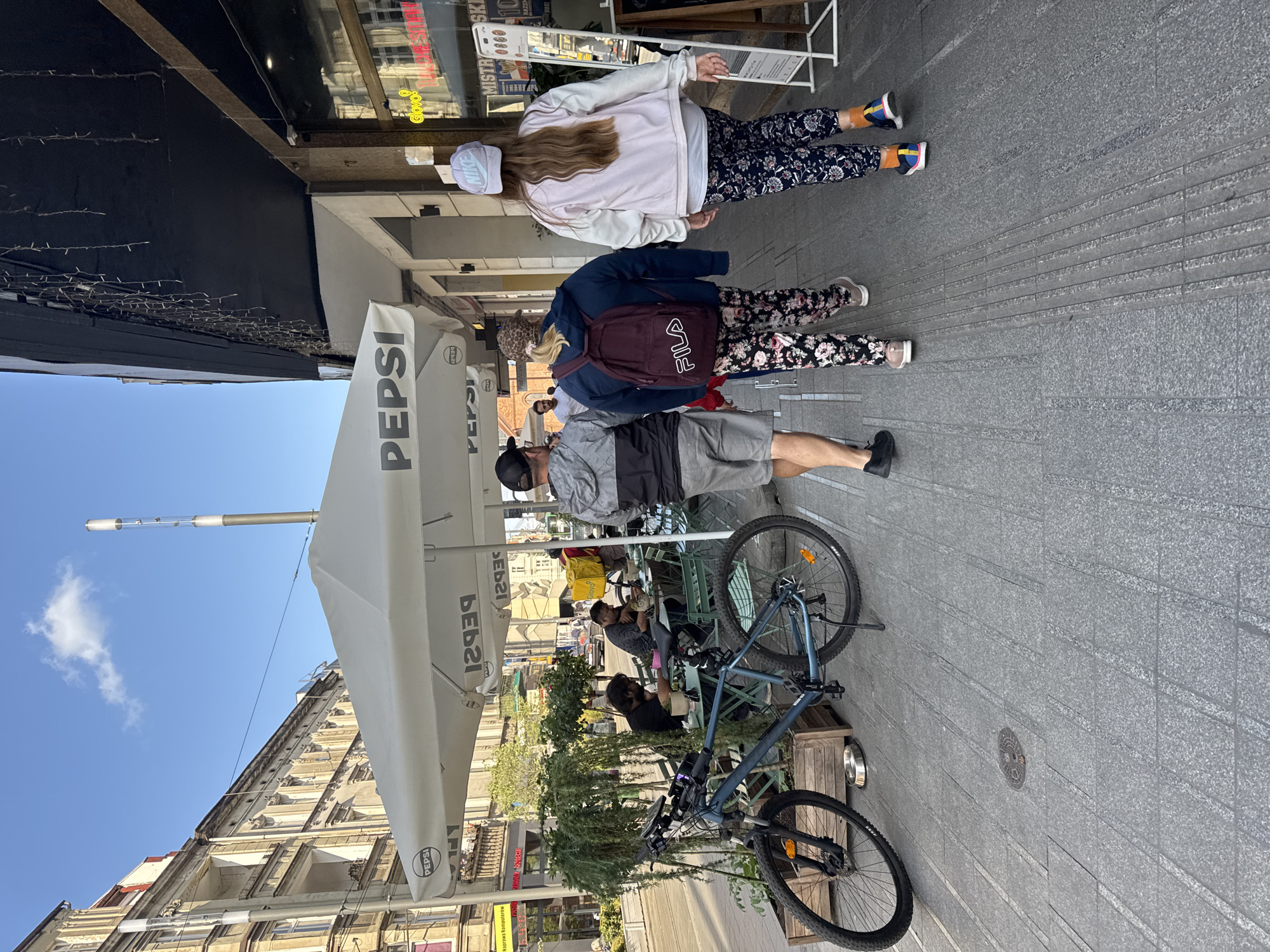 The image depicts a sunny day on a city sidewalk in Poznań, Poland, likely in the Old Market Square area due to the architecture and outdoor seating. In the foreground, a blue bicycle with a phone mount is parked next to a planter. Three people are walking away from the camera on the sidewalk. The person on the left is a male wearing a gray jacket and shorts. The middle person is wearing a navy hoodie and floral patterned pants, carrying a maroon backpack with "FILA" printed on it. The person on the right is wearing a light pink jacket and dark floral patterned pants, with a light blue "NIKE" cap.
Further back, under a white umbrella with "PEPSI" branding, two men are seated at tables with green chairs. One man is wearing a black t-shirt and the other a dark gray t-shirt. The background shows buildings with classical architecture and a street with traffic. The overall scene is a typical urban environment with people enjoying the outdoors and a beverage.