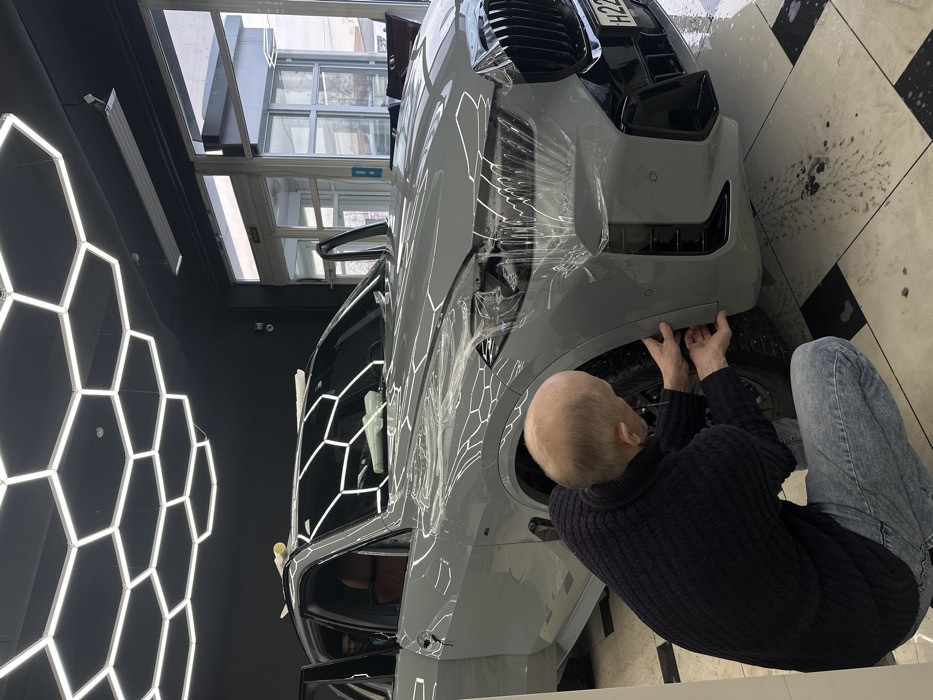 The image depicts an indoor scene within an automotive detailing facility in Saint Petersburg, Russia, as evidenced by the '78 RUS' region code on the vehicle's front license plate. A light gray BMW X5 SUV is positioned centrally, undergoing the application of clear paint protection film. The front bumper, headlights, and driver's side front fender are partially covered with the wet, transparent film.
A bald male individual, wearing a dark blue ribbed sweater and light blue jeans, is crouched beside the driver's side front fender, actively manipulating the protective film with both hands near the wheel arch. Water or an application solution is visible on the vehicle's surface and dripping onto the black and white checkered floor tiles. The driver's side front door is ajar, revealing a reddish-brown leather interior and the partial silhouette of a second individual with dark hair observing from within the vehicle.
The facility features a dark ceiling equipped with hexagonal LED light fixtures, which are prominently reflected on the car's glossy surfaces and the wet floor. In the background, large glass windows and an automatic sliding glass door lead to an exterior view under an overcast sky. A white industrial heater is mounted on the ceiling above the windows. Rolls of film or paper are resting on the vehicle's roof. The visible front license plate reads 'H227XX 78 RUS'.