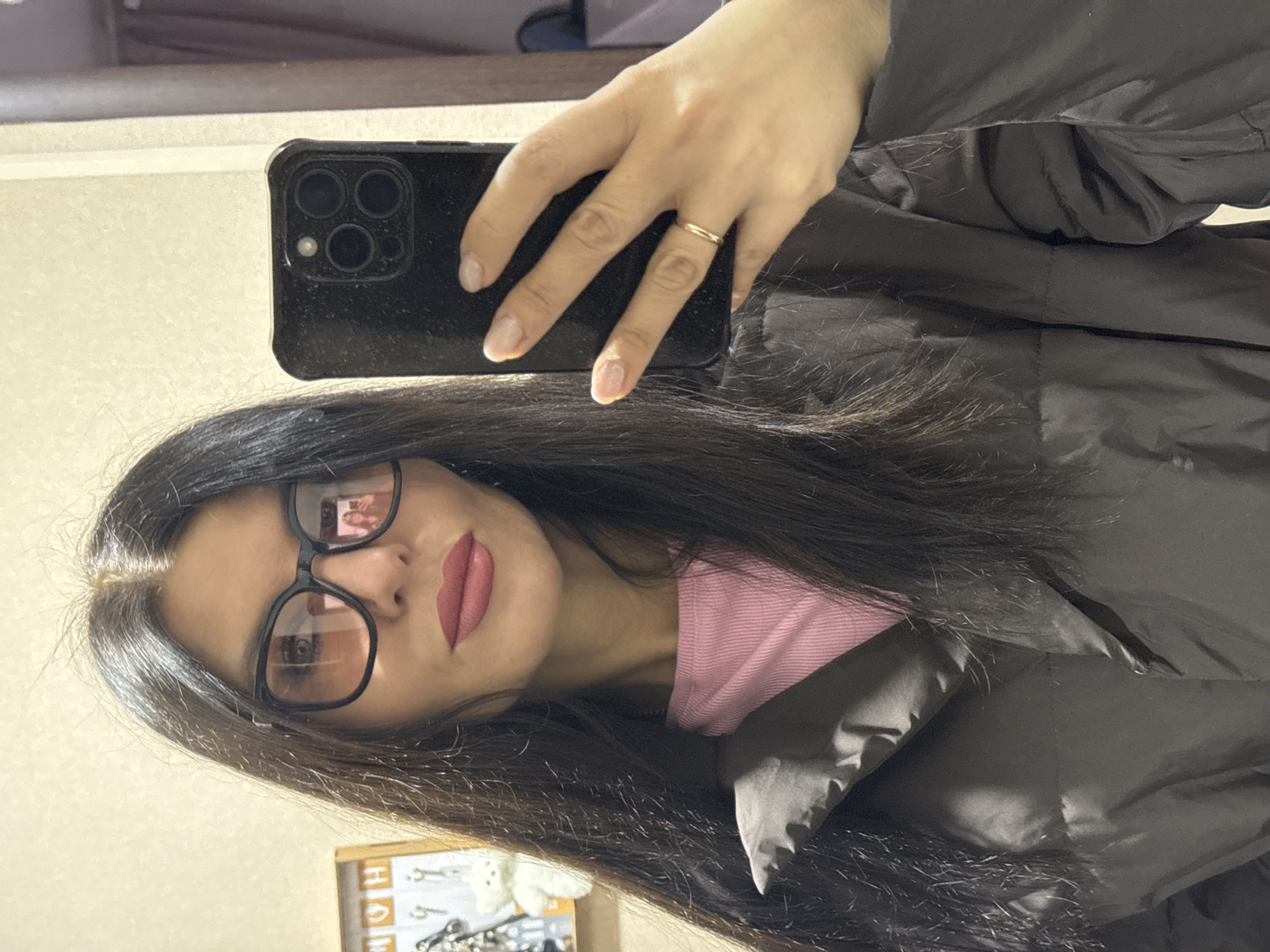 A woman in glasses takes a selfie with her phone. She is wearing a pink shirt and a black puffer jacket. She has dark hair and is wearing lipstick. The background appears to be a room, potentially located in Chaikovsky, Russia, although this is based only on the implicit information of the source of the image. The reflection in her glasses suggests there is another person in the room.