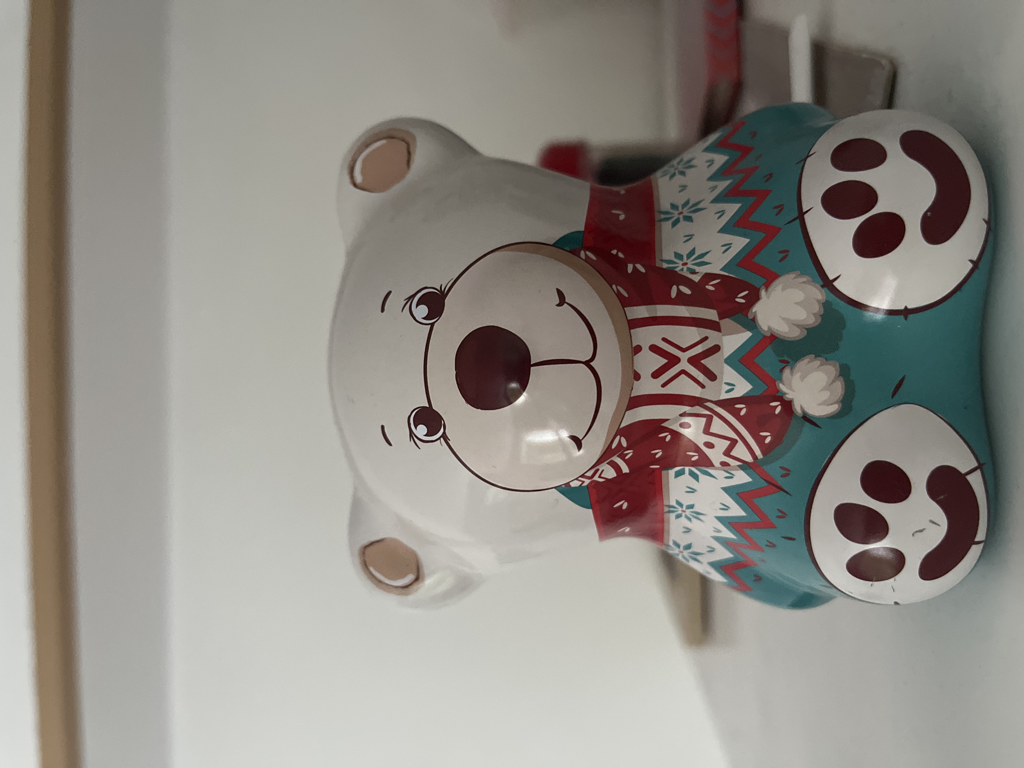 A white, ceramic bear-shaped object, decorated with a red and white Christmas-themed sweater and a teal base, sits on a white surface. The bear has a large, dark red nose, friendly eyes, and a smiling mouth. Its sweater features a snowflake pattern and a red zigzag trim. The teal base is adorned with white paw prints, each containing a smiling mouth. The background is a plain white wall, with a light wooden surface visible at the top. The scene appears to be indoors, possibly on a shelf or table. There is no discernible connection to the city of Kohtla-Järve linn, Estonia.