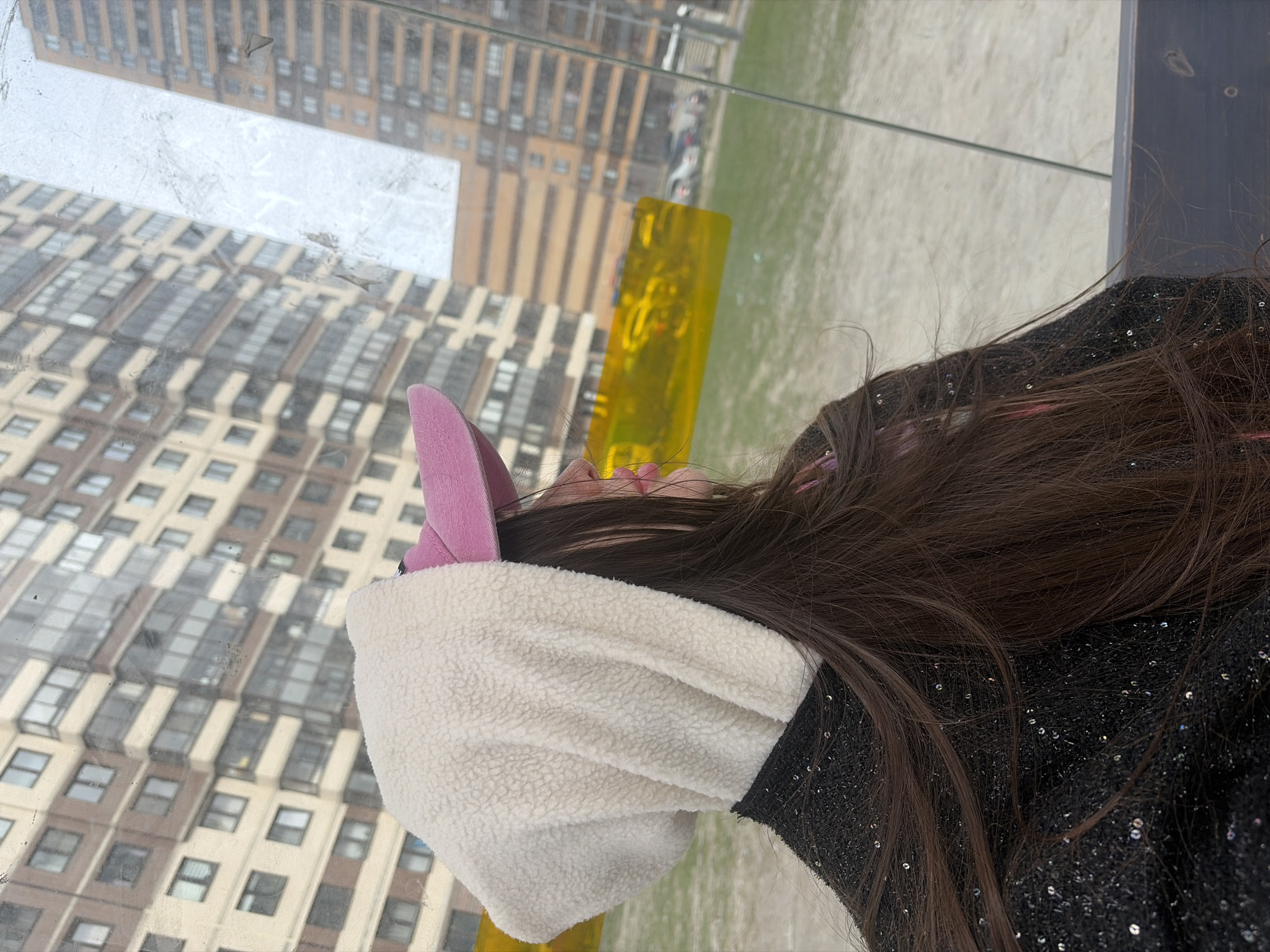 The image captures a person from a side/back perspective, looking out through a large, slightly smudged glass window. The individual has long, dark brown hair and is wearing a white, fluffy hood with distinctive pink, pointed ears, reminiscent of an animal. A dark garment, possibly black, with shimmering sequins is visible on their upper back.
Beyond the window, an urban cityscape unfolds. Modern, multi-story buildings with numerous windows, displaying brown and beige facades, stretch across the background. In the mid-ground, a striking, bright yellow, rectangular structure or art installation is situated on a verdant, grassy expanse. The lighting indicates it is daytime, likely an overcast day, due to the soft, diffused illumination and absence of strong shadows. The overall scene suggests a moment of contemplative observation within an urban environment. This setting is located in Saint Petersburg, Russia. No visible text is present in the image.
