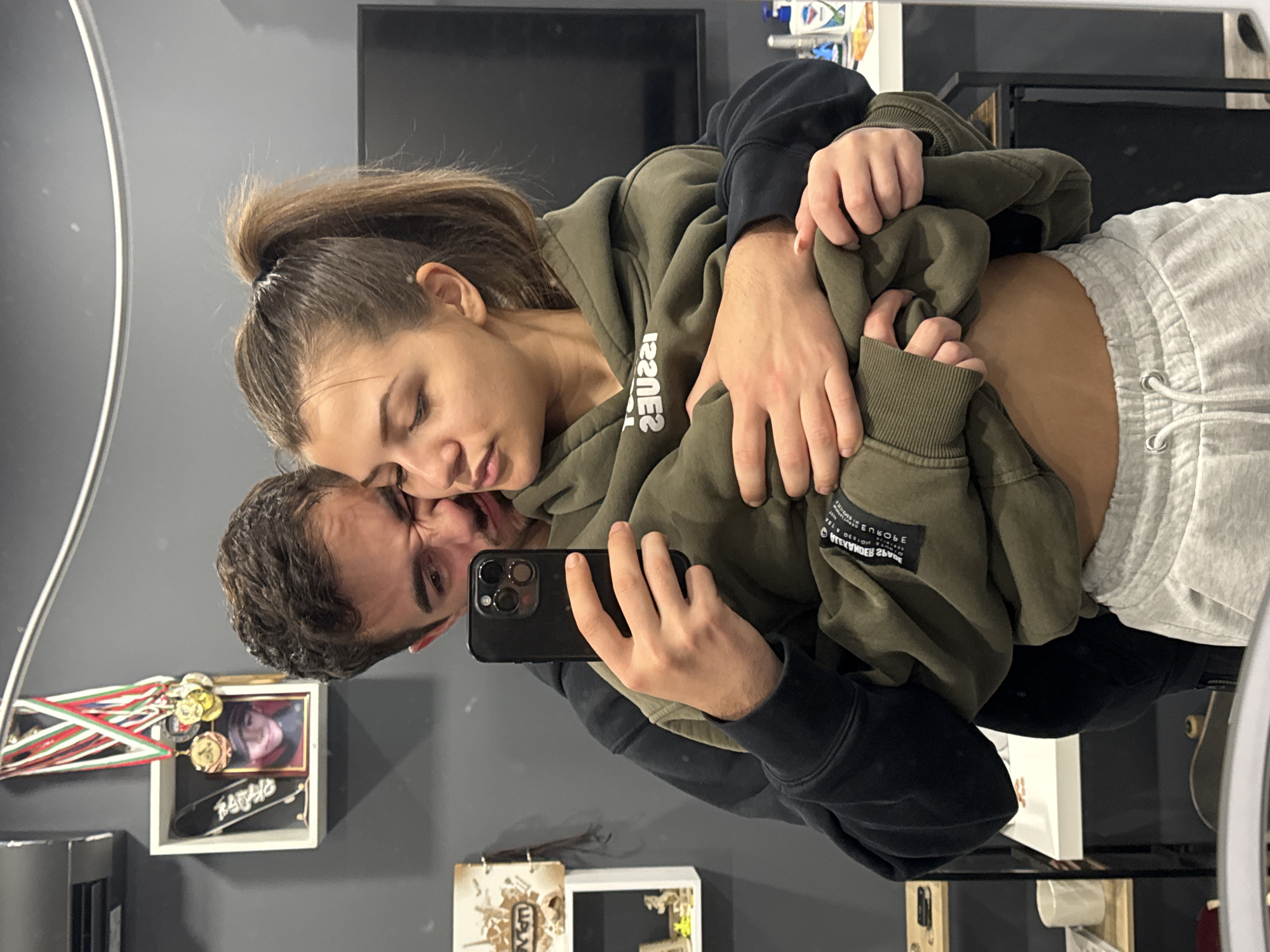 A young man and woman are posing for a mirror selfie. The man is holding a black smartphone with three camera lenses. He is looking at the camera with a playful facial expression. He wears a black hooded sweatshirt with an olive green hooded sweatshirt partially covering his torso. The woman is leaning her head on the man, appearing relaxed with her eyes closed. She has her hair pulled back in a ponytail and is wearing the olive green hooded sweatshirt. A portion of her midriff and grey sweatpants is visible. The background shows a grey wall, and several objects including a framed photo display with medals, a small skateboard, a television and a shelf holding various items. The image appears to have been taken indoors. The location is indeterminable, with no explicit indication of Sofia, Bulgaria in the scene.