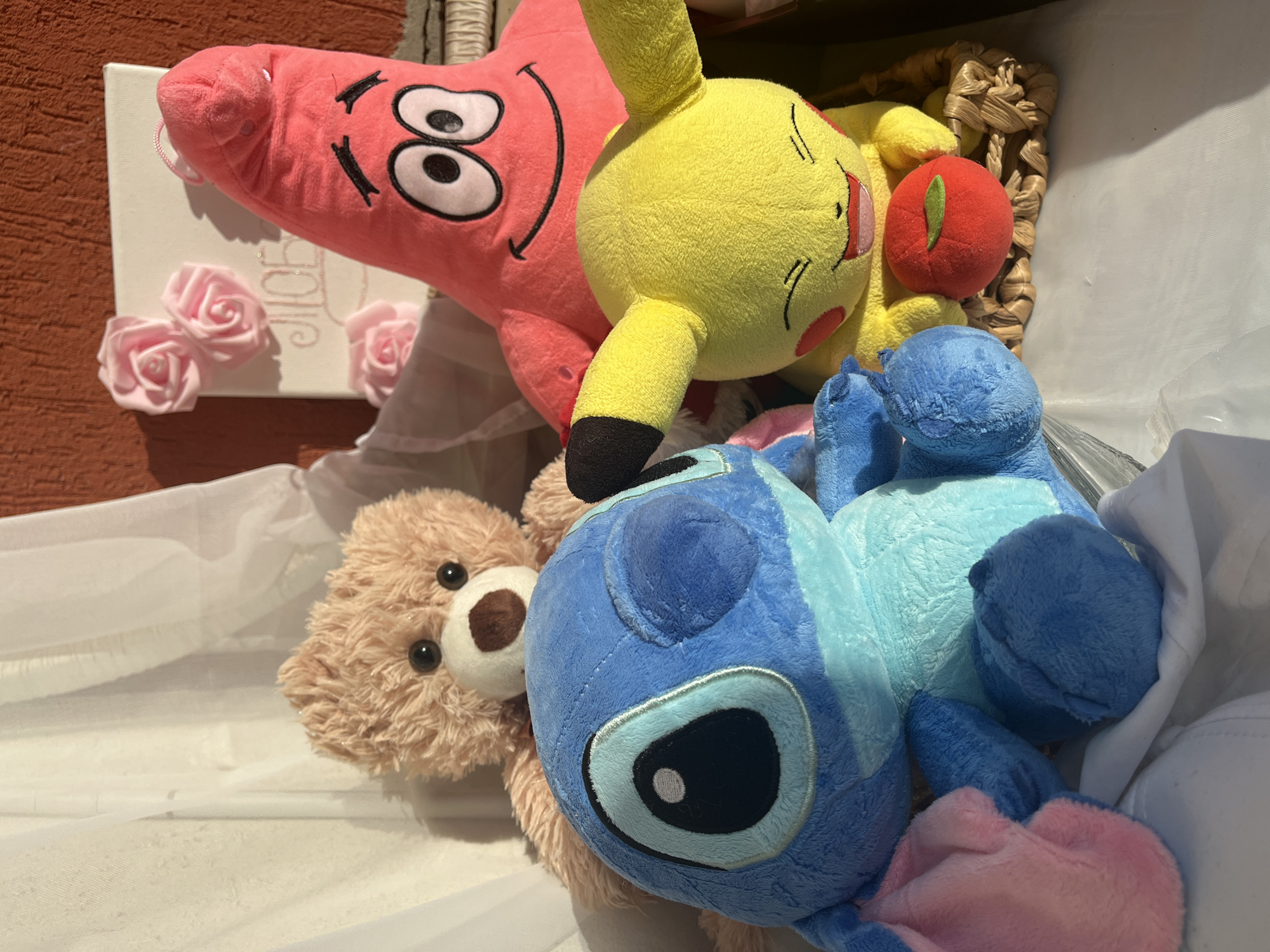 A close-up, eye-level shot of a collection of plush toys, including a pink Patrick Star, a yellow Pikachu, a blue Stitch, and a brown teddy bear, arranged together. The toys are lying on a white cloth with a textured surface. A woven basket is visible in the background, slightly obscured by the toys. A white canvas decorated with pink fabric roses is seen partially in the upper-left, resting against a textured orange wall. The location is specified as Veliko Tarnovo, Bulgaria, though there are no unique indicators of this.