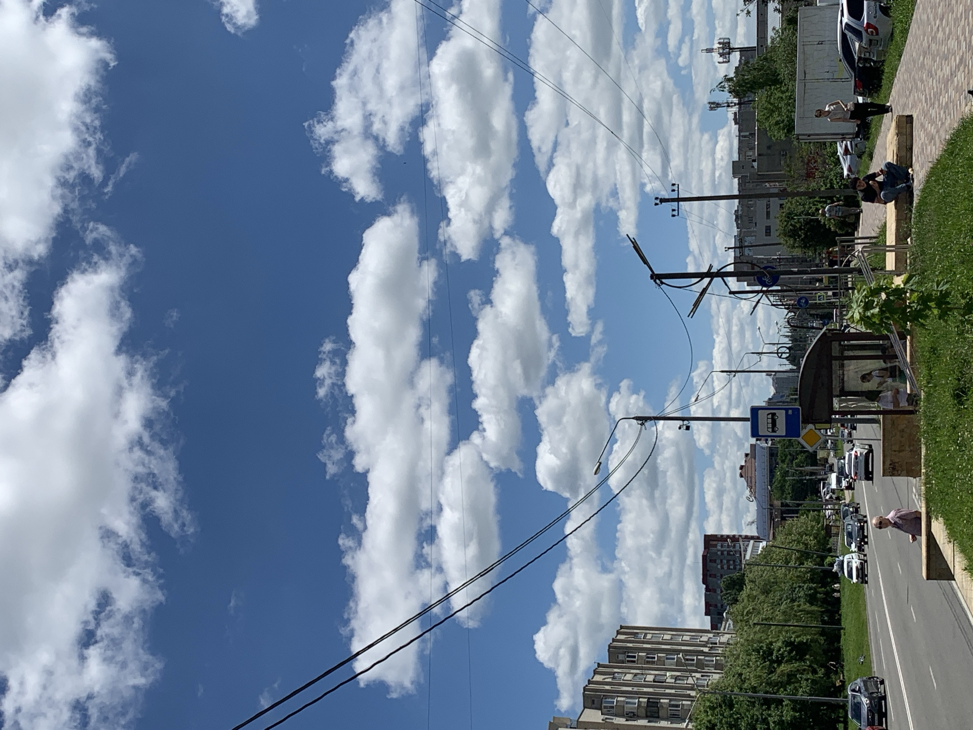 In an urban setting, a street runs horizontally across the lower portion of the image, with a sidewalk and grassy area to the right. The street is lined with vehicles, including cars and a bus. Several people are visible on the sidewalk and by the bus stop, some sitting and others standing. Buildings, including apartment complexes and what appear to be commercial structures, form the backdrop. The sky is a vivid blue, filled with fluffy white clouds. Power lines and streetlights are prominent overhead, crisscrossing the frame. The scene appears to be a typical daytime street view in a city.