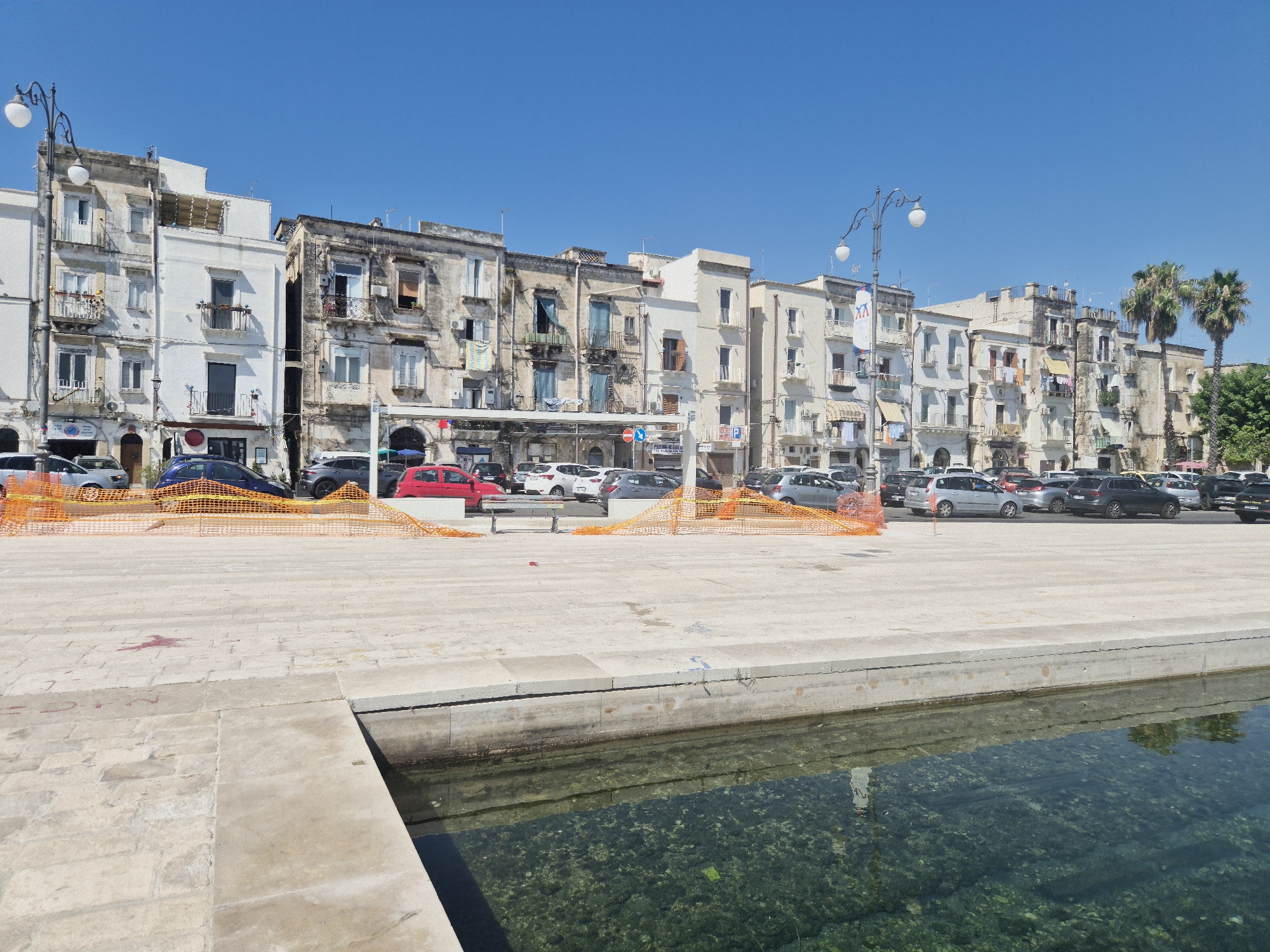 This is a wide shot of a waterfront promenade in Taranto, Italy. In the foreground, a concrete promenade borders a clear body of water, revealing submerged rocks. Beyond the promenade, a line of older, multi-story buildings with balconies and weathered facades stretches along a street filled with parked cars. A distinctive lamppost with two globes stands on the left side of the frame. Two palm trees are visible to the right, with dense green foliage behind them. The sky is a clear, bright blue. Orange construction netting is present in front of the buildings, separating the promenade from the street. The overall scene depicts a sunny day in a coastal urban environment.