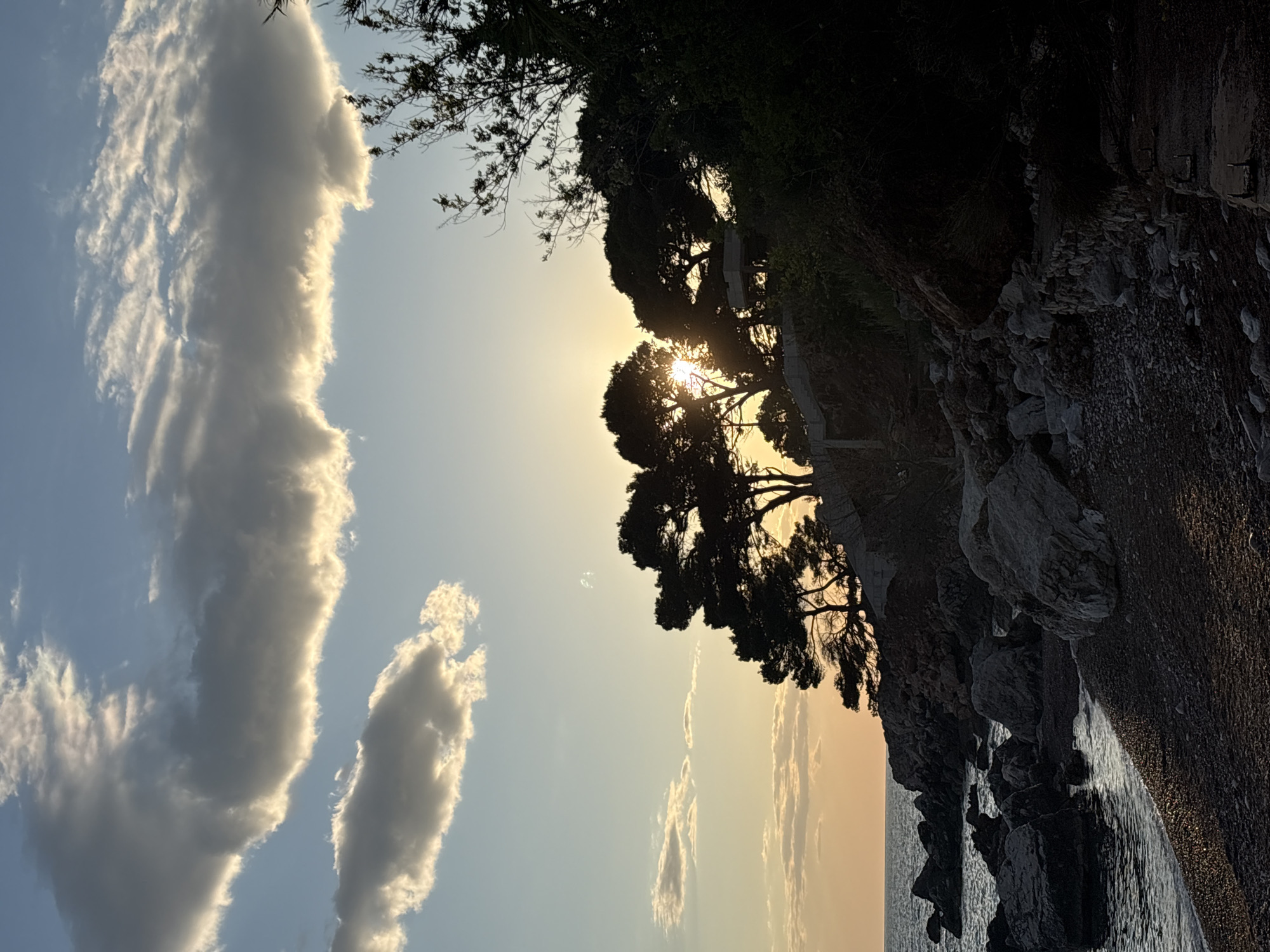This landscape photograph, taken in Bar, Montenegro, captures a serene coastal scene during what appears to be sunset or sunrise. The sky dominates the upper and left portions of the image, showcasing a gradient from a soft blue at the top to warmer yellow and orange hues closer to the horizon on the right.
Prominent in the sky are several clouds, most notably a large, elongated cloud formation that stretches vertically on the left side, illuminated from behind by the sun, creating bright, glowing edges. Smaller, scattered clouds are also visible.
On the right side of the image, the sun is partially visible, peeking through the dense foliage of silhouetted trees, likely pines or similar evergreens, growing along a rocky incline. The strong backlight from the sun turns the trees into dark, intricate silhouettes. Below these trees and rocks, the calm waters of the sea are visible, reflecting the warm light of the sky. The shoreline consists of dark, rugged rocks and what appears to be a pebbly or sandy beach in shadow.
No people or visible text are present. The overall atmosphere is peaceful and natural, emphasizing the beauty of the coastal landscape at golden hour.