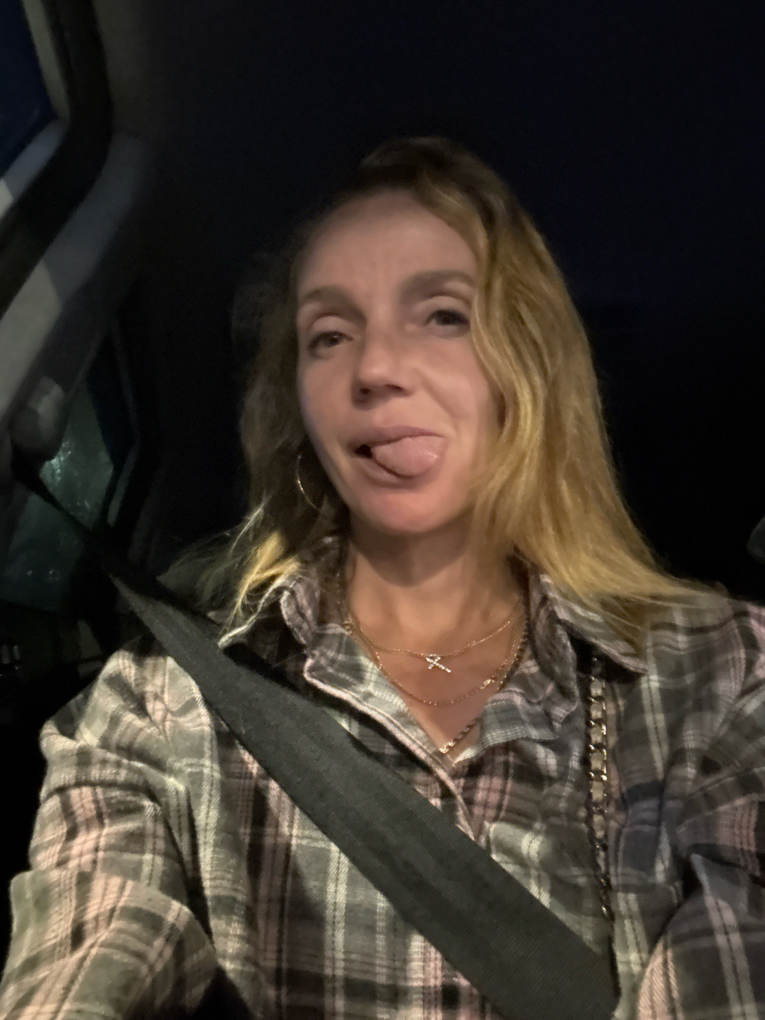 A woman with blonde hair is in the driver's seat of a car, with her seatbelt fastened. She is wearing a grey and pink plaid shirt and multiple necklaces. Her tongue is sticking out, and she is looking at the camera. The background is dark, suggesting it is nighttime or inside a dark environment. There is no visible indication of the city of Saint Petersburg, Russia.