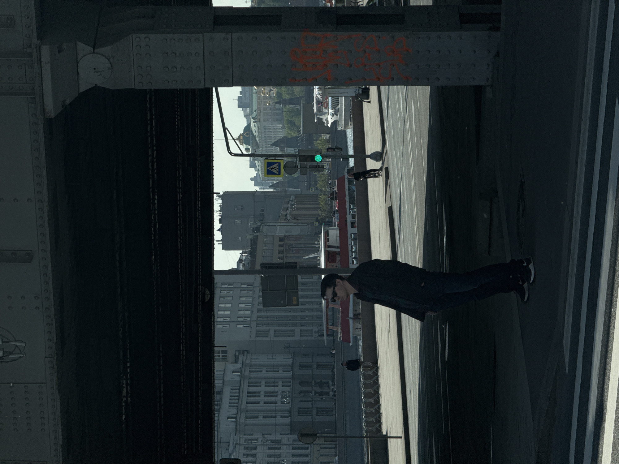 The image shows a view under a bridge in Moscow, Russia. The view is framed by the bridge's steel structure, with rivets and graffiti visible on the metal. In the background is a cityscape with a mix of buildings. A traffic light shows a green light. A pedestrian crossing sign is also visible. A man wearing dark clothing and sunglasses is walking on the sidewalk. Other people can be seen in the distance near the traffic light.