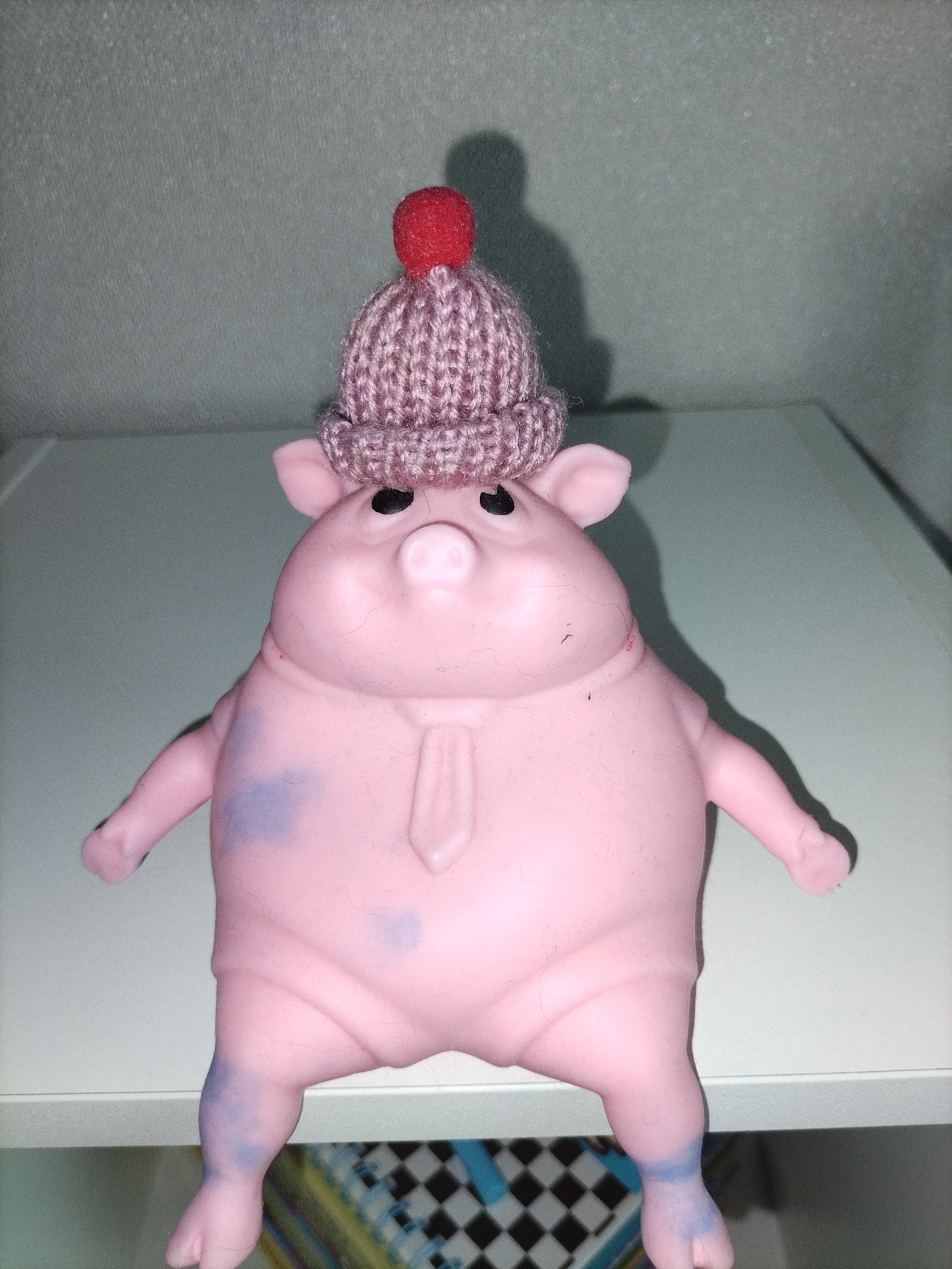 A close-up, indoor photograph features a pink, pig-shaped toy or figurine standing upright. The toy is made of a soft, possibly rubber or plastic, material and has a generally plump body with small, stubby arms and legs. It wears a small, knitted, light purple or mauve beanie with a prominent red pom-pom on top of its head. The pig has black, round eyes and a sculpted tie molded onto its chest. There are noticeable blueish-purple stains or discolourations on its left side/arm and both legs.
The pig stands on a smooth, light-colored surface, possibly a shelf or tabletop. A distinct shadow of the pig and its hat is cast on the light grey wall behind it, suggesting a light source from the upper right. Below the surface the pig is standing on, a partial view of a checkered pattern (black and white squares) and some colorful elements are visible, suggesting other items on a lower shelf or in the background. The overall setting is indoors, with no specific time of day discernible. No text is visible in the image.