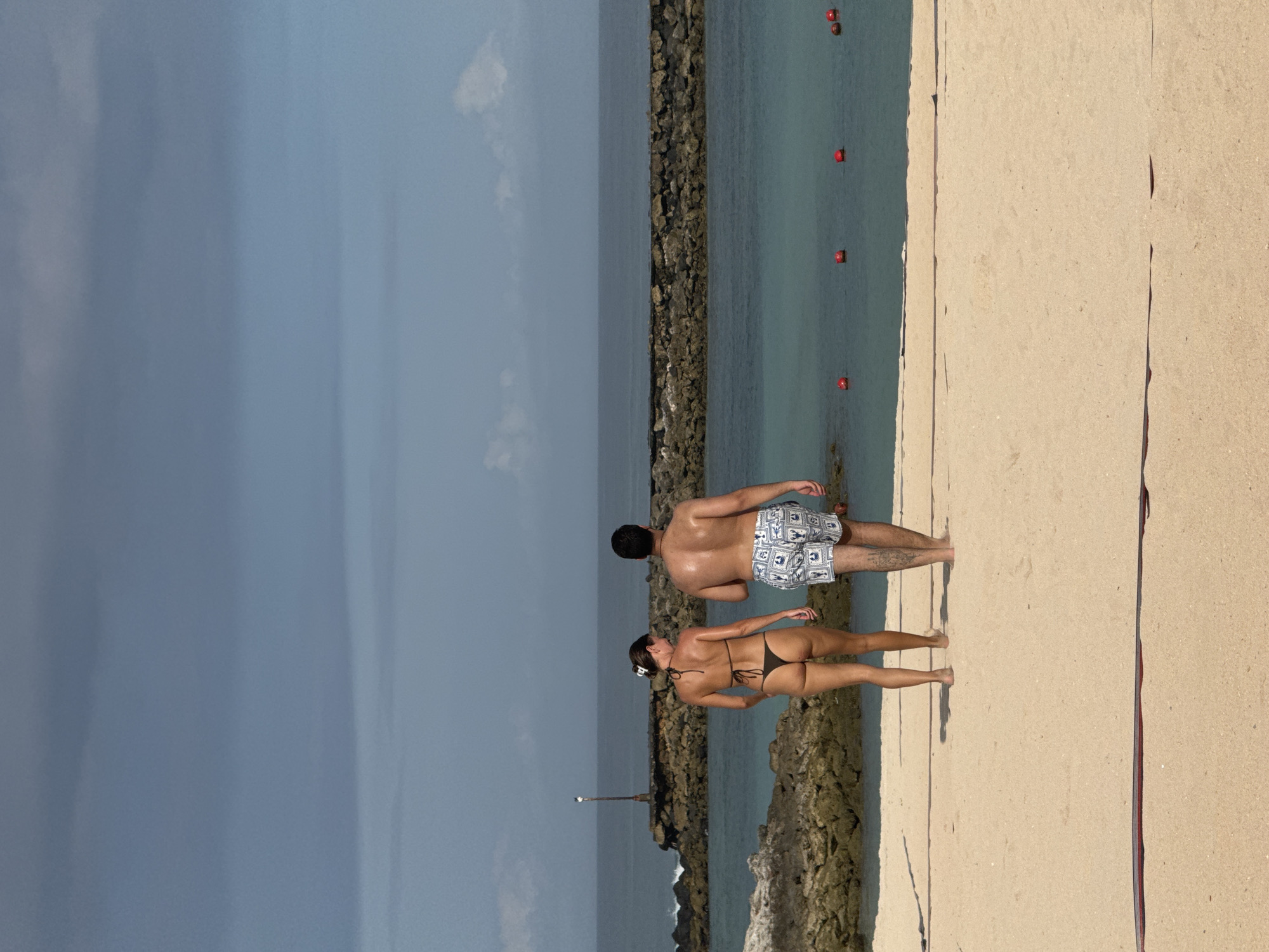 The image captures a man and a woman standing at the edge of a beach where it meets a rocky breakwater, the location is in the city of Kuta Selatan, Indonesia. The man is wearing patterned swim trunks, and the woman is wearing a dark bikini. They are both facing the water, which extends to the horizon where it meets the sky. The sky is partly cloudy with a mix of blue and gray hues. Red markers are visible in the water, indicating some form of boundary or marking. The sand is a light tan color, and a darker colored object is visible on the beach in the right side of the frame. A small metal pole is on the breakwater.