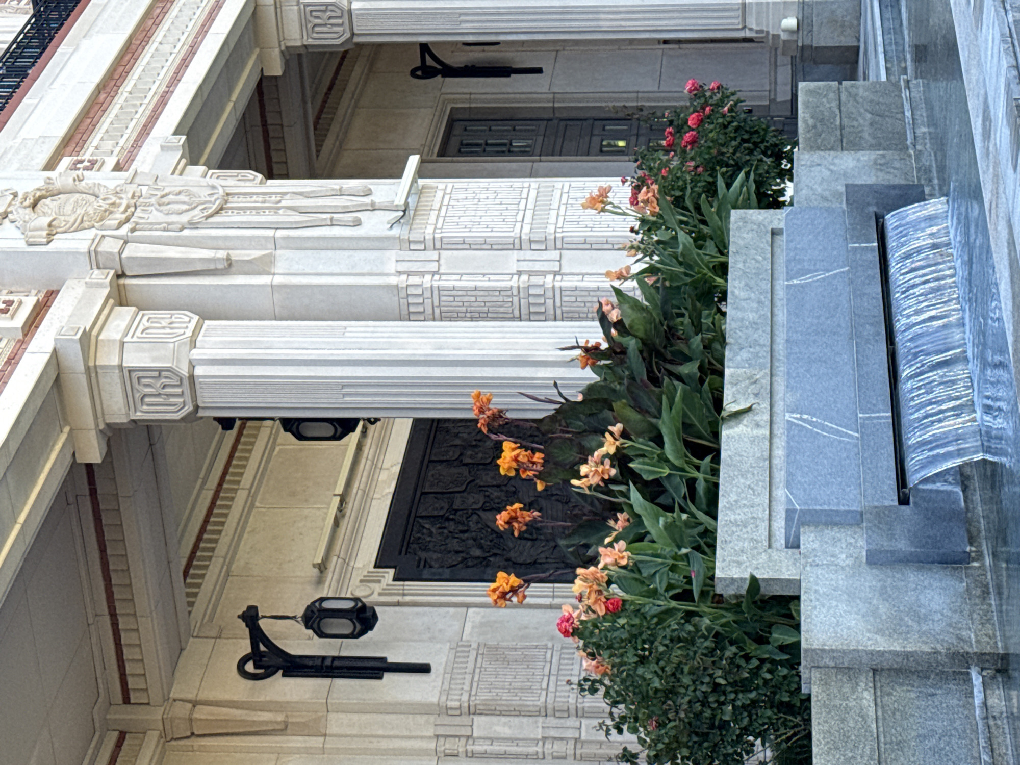 The image shows the facade of a building with classical architectural elements. There are fluted columns, decorative carvings, and ornate metal lanterns mounted on the exterior walls. In front of the building, there is a tiered planter filled with lush green foliage and vibrant orange and pink flowers. A modern-style water feature with cascading water is visible in the foreground. The overall impression is one of grandeur and elegance. The location is the Palace of Agriculture in Sevastopol, Ukraine.