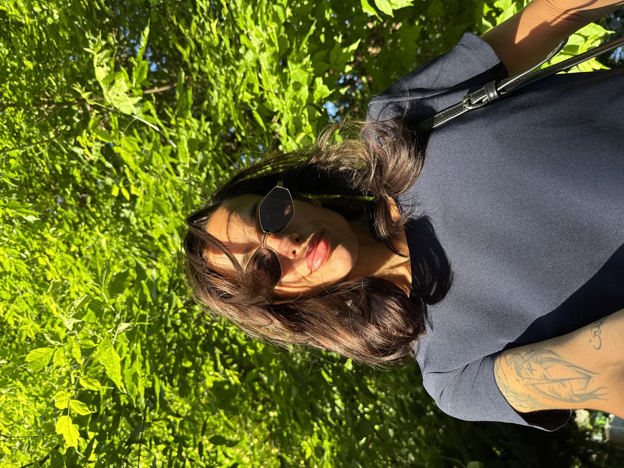 A medium shot captures a woman with brown hair and sunglasses, posed against a background of lush green foliage, likely taken outdoors in Pleven, Bulgaria. She wears a dark-colored top and has a visible tattoo on her arm. A black strap, possibly from a bag, is visible across her shoulder. Sunlight filters through the leaves, creating dappled lighting on her face and the surrounding vegetation.