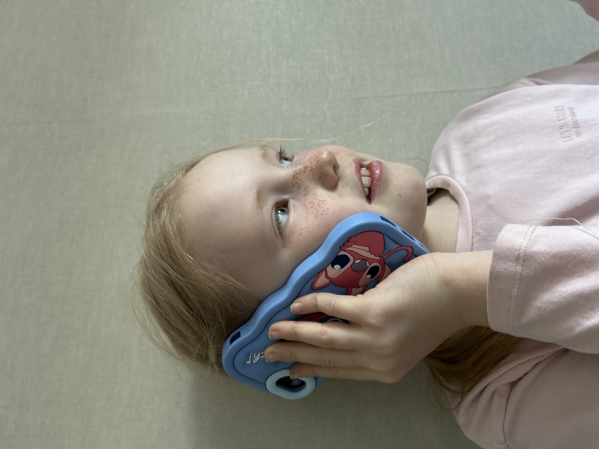 A fair-skinned child with freckles and blonde hair is lying on a light beige surface. The child is wearing a light pink, long-sleeved shirt with the words "LET'S START YOU CAN DO IT" visible on the shirt. The child is holding a light blue cell phone in their right hand, with a pink cartoon character on the phone case. The child is smiling and looking upward. The location is Chaikovsky, Russia.